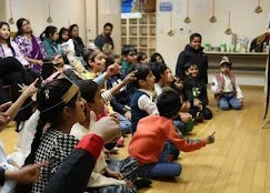 Group of children and adults sitting on the floor in a classroom, engaging in an activity.