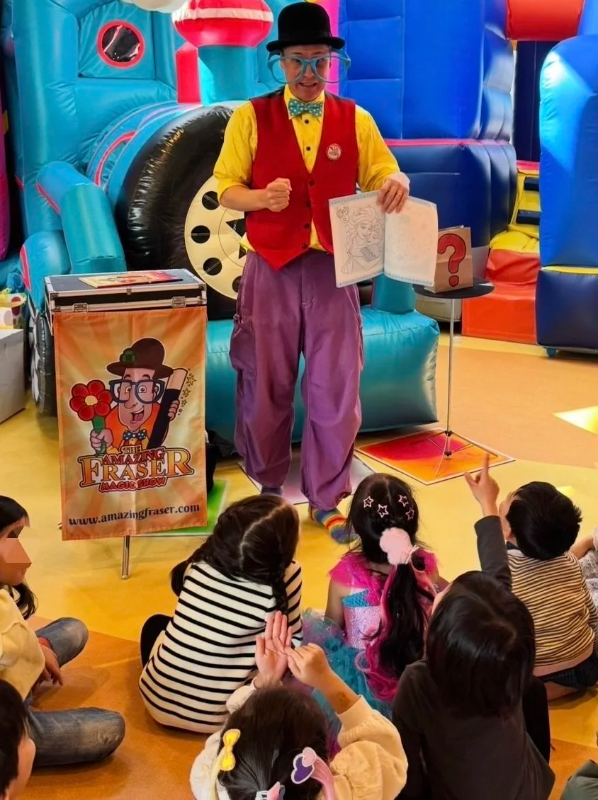 A clown in a yellow shirt, red vest, purple pants, and a black bowler hat performing a magic show for children in a colorful indoor play area. The clown is holding a drawing and standing next to a box with a cartoon clown logo.