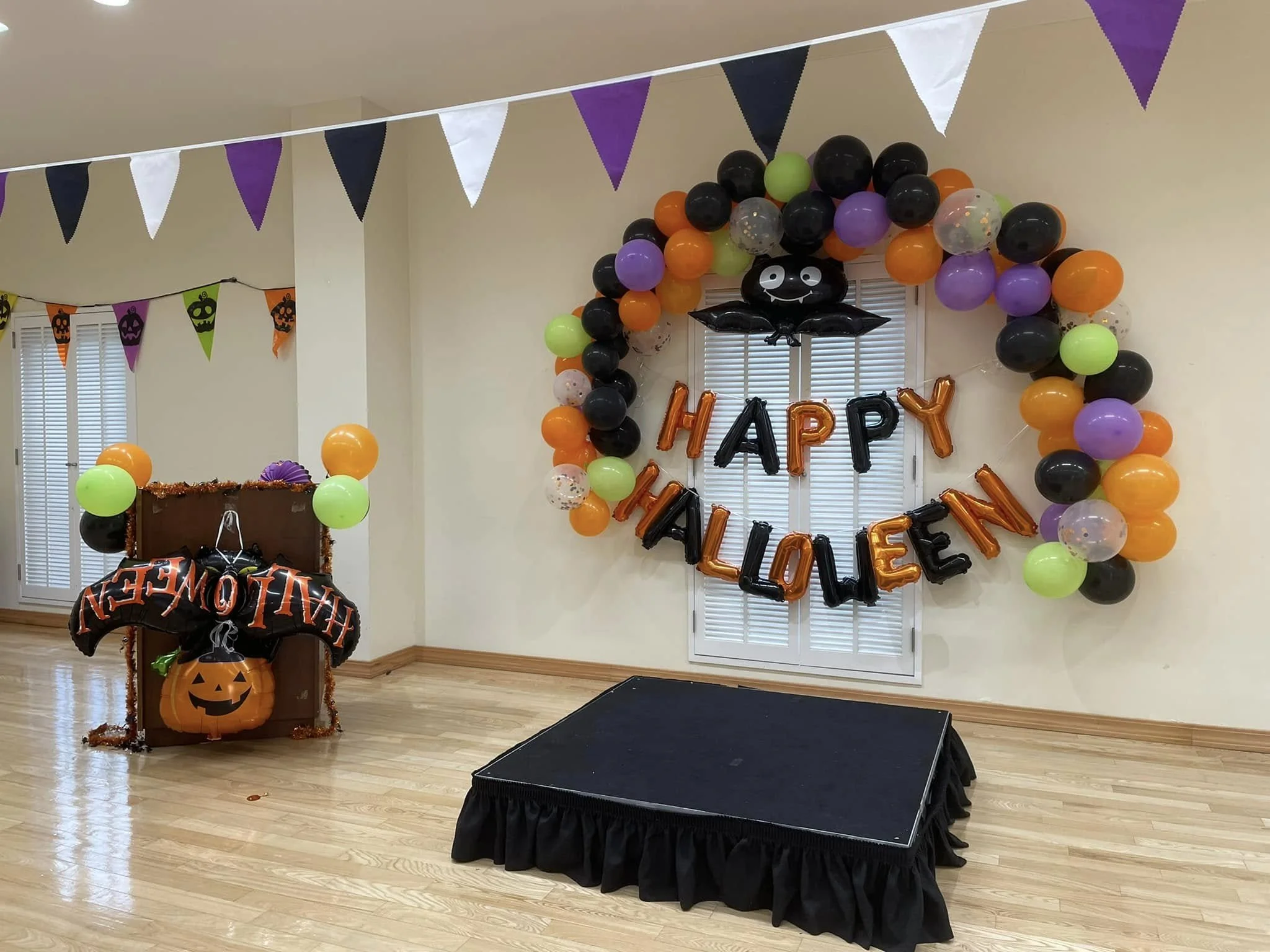 Halloween party decorations with as a balloon arch featuring black, orange, purple, and green balloons, a pumpkin scarecrow, and banners with pumpkin faces, with a saying 'Happy Halloween' in balloons.