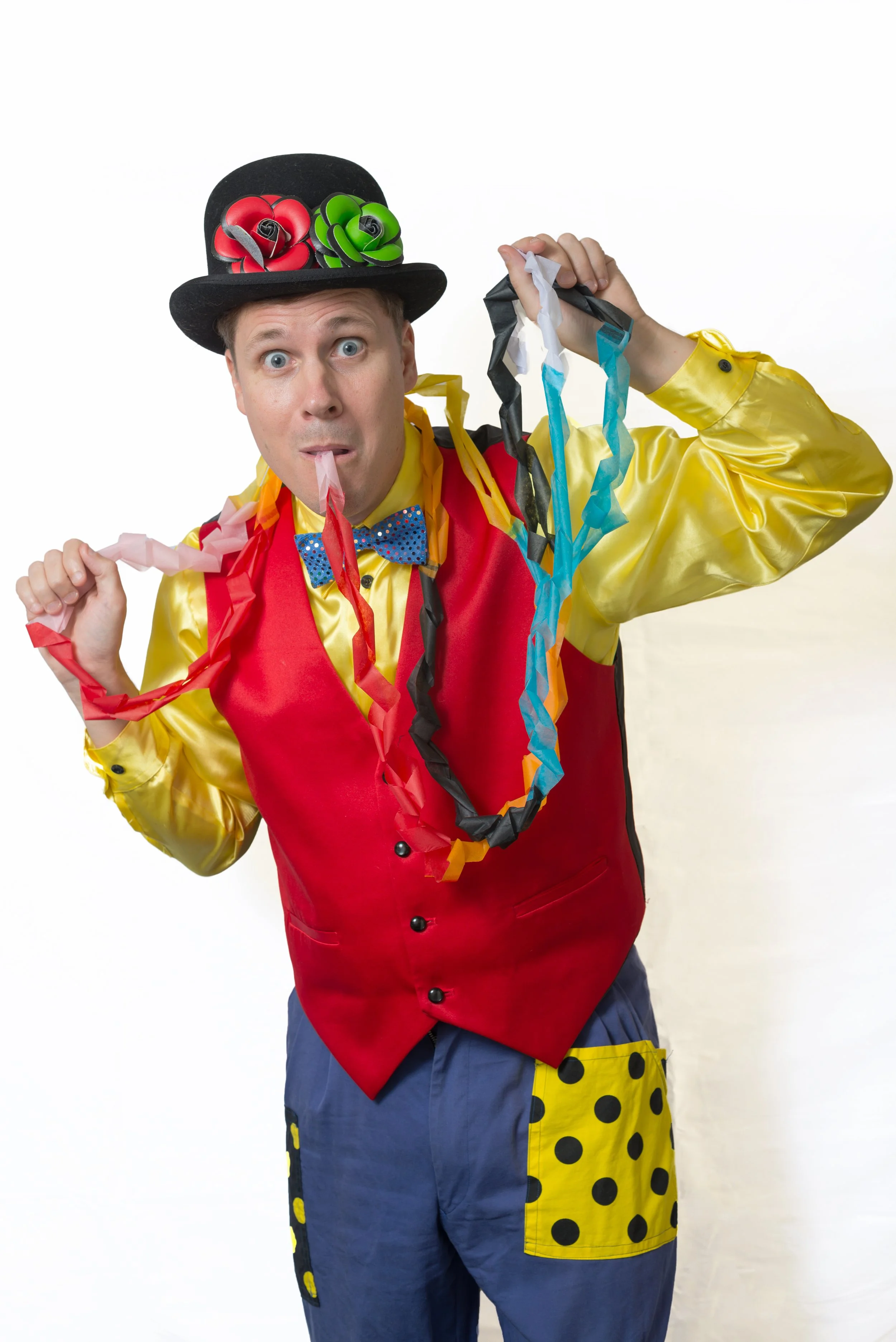 A man dressed as a clown wearing a black top hat with red and green pinned fabric flowers, a yellow shirt, a red vest, and striped blue and yellow clown pants, holding colorful streamers and sticking out his tongue.