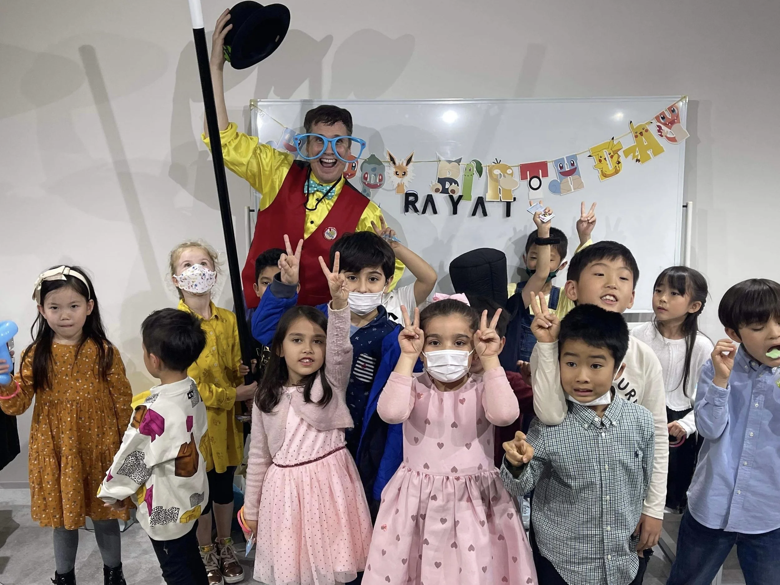A group of children posing with a clown or entertainer. The children are making peace signs and some are wearing masks. The clown is wearing large glasses, a yellow outfit, and a red vest. Behind them is a whiteboard with a colorful banner that has c