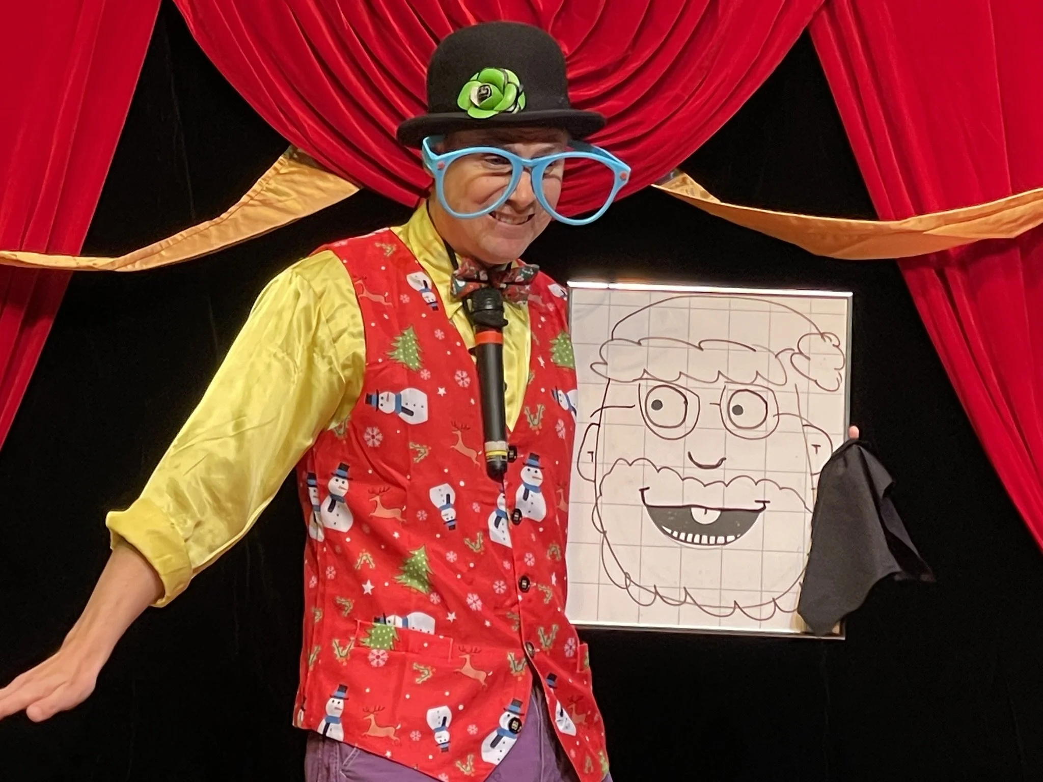 A man wearing large blue glasses, a black top hat with a green flower, a yellow shirt, and a red holiday vest with snowmen, standing in front of red curtains with gold trim, holding a drawing of a happy face.