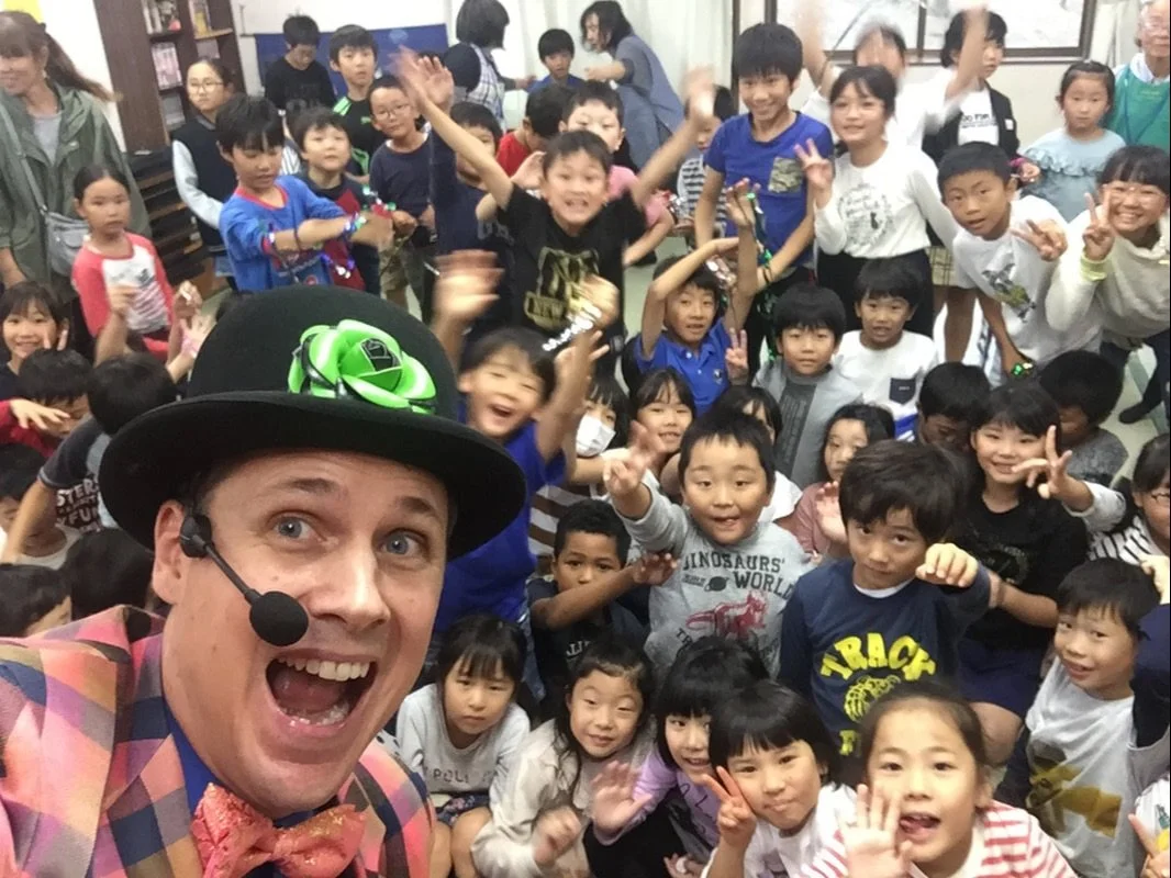 The magician 'The Amazing Fraser' wearing a colorful suit, bow tie, and a black top hat with a green decoration, taking a selfie with a large group of children in a classroom or event setting.