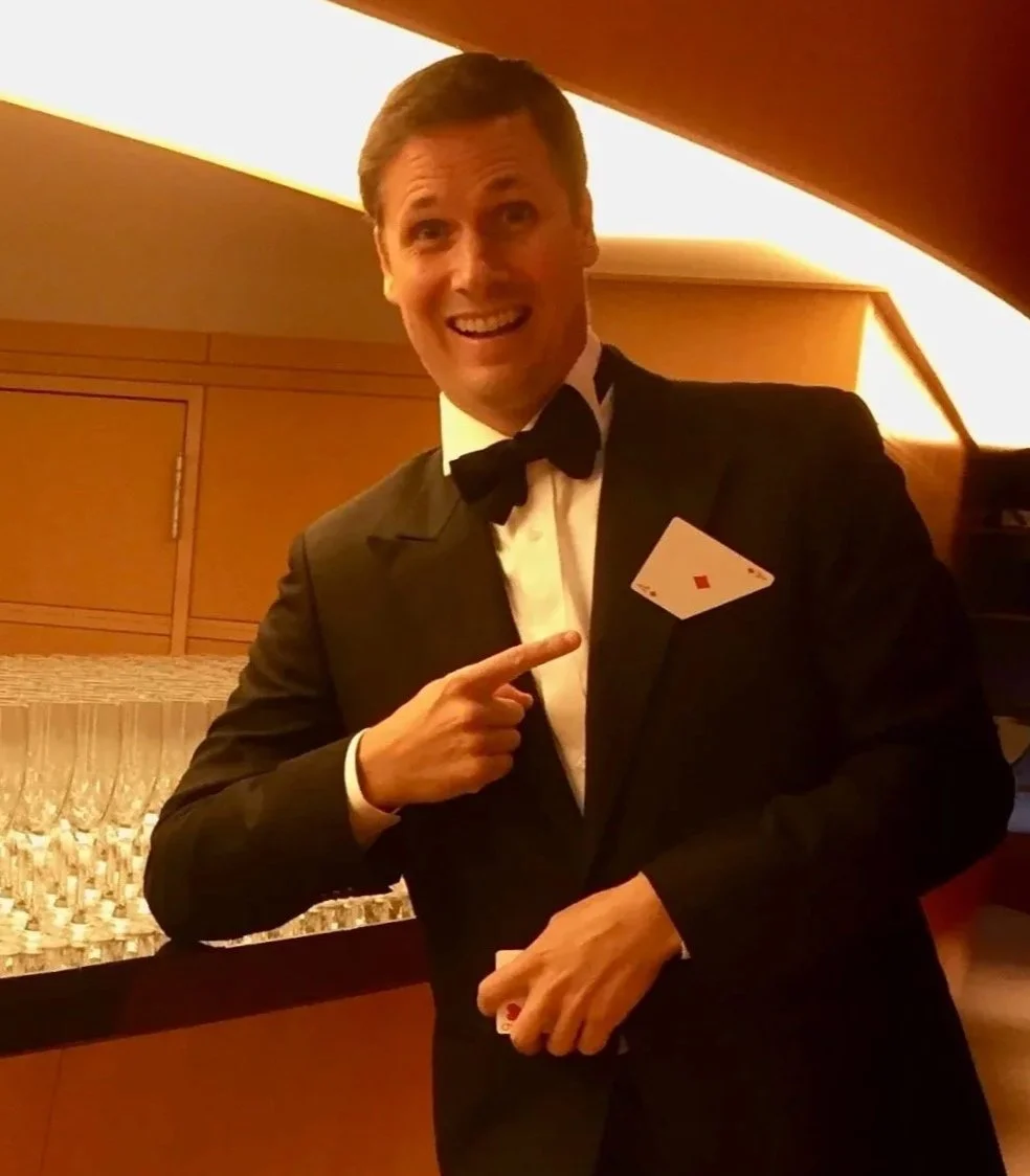 Magician in a tuxedo with a bow tie, pointing at a playing card (ace of diamonds) tucked into his tuxedo pocket, smiling and standing in a warm-lit room.