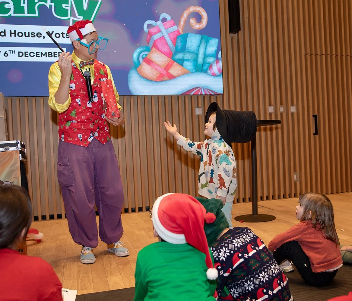 A clown in a colorful costume and large glasses is entertaining children during a Christmas event. A young boy is raising his hand to ask a question, while other children sit on the floor, with some wearing Christmas hats and sweaters. There is a lar