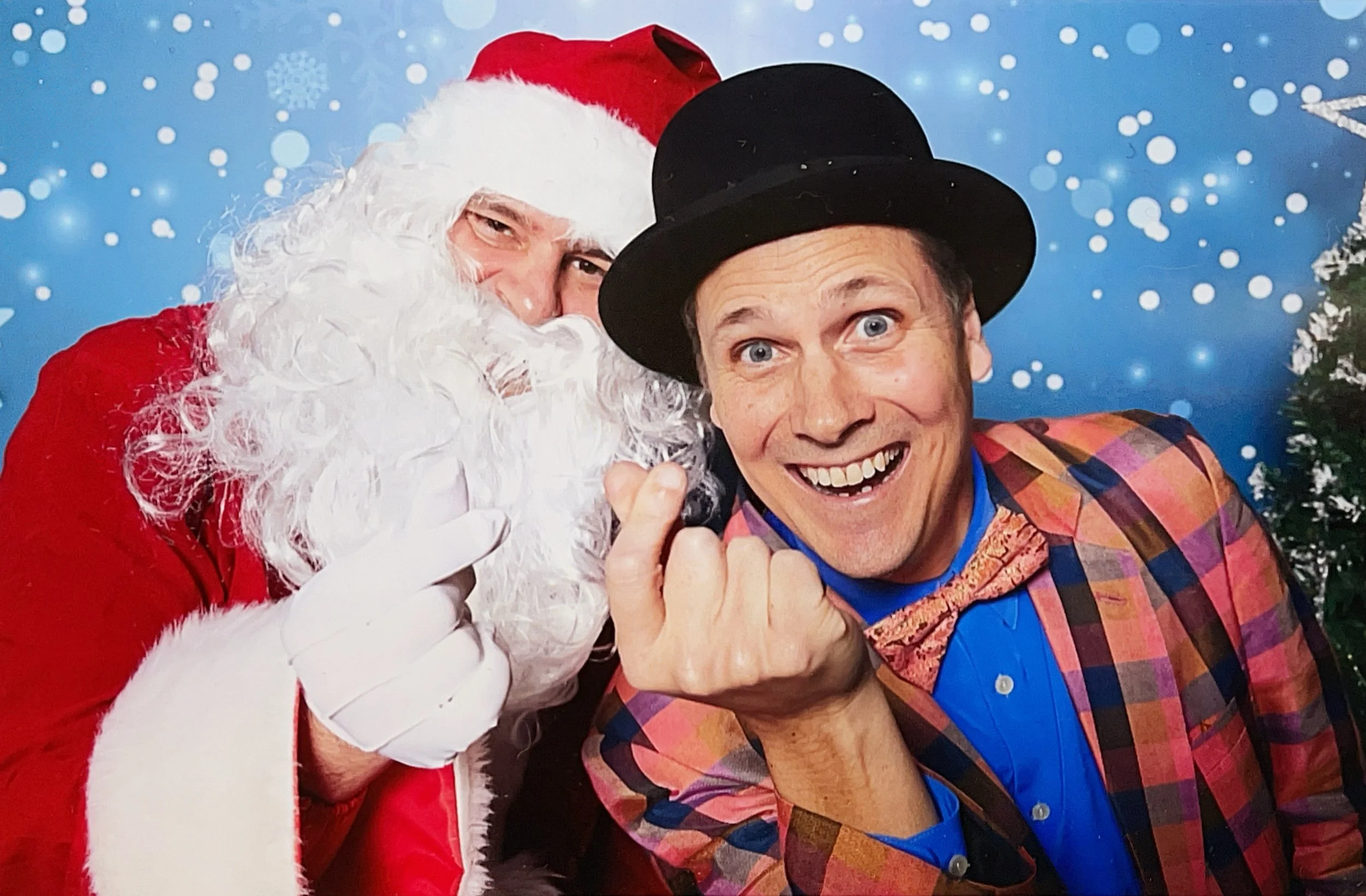 Santa Claus with a white beard and red suit paired with the magician The Amazing Fraser. They are smiling together in front of a snowy, festive background.
