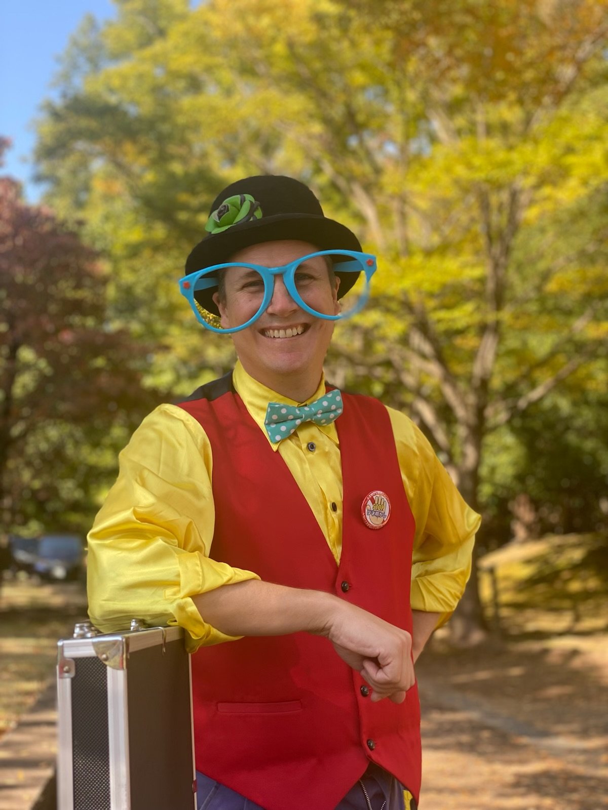 Kidshow magician The Amazing Fraser dressed as a clown or performer outdoors in front of trees with autumn foliage, wearing large blue glasses, a black hat with a green flower, a red vest over a yellow shirt, a polka dot bow tie, and a badge.