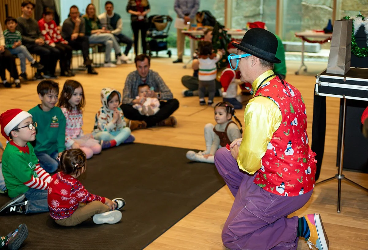 A clown wearing colorful clothing, including a vest with snowmen and Christmas trees, yellow shirt, purple pants, and a black hat, performing for children at a Christmas party. The children are sitting on a black mat, wearing festive sweaters and hat