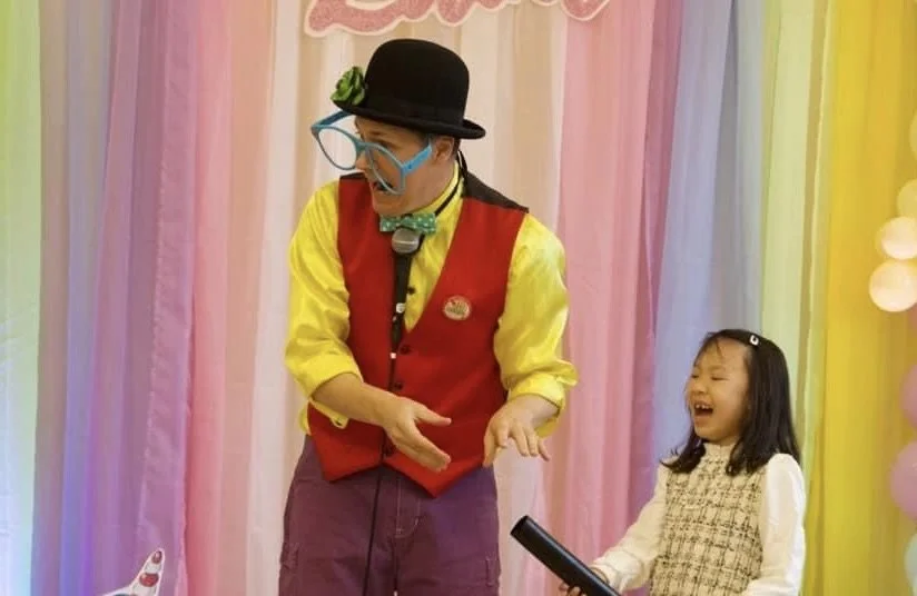 A clown dressed in a yellow shirt, red vest, and purple pants, wearing a black hat, glasses, a green bow tie, and a mask, entertaining a young girl with a joyful expression at a colorful event with rainbow-colored curtains and balloons.