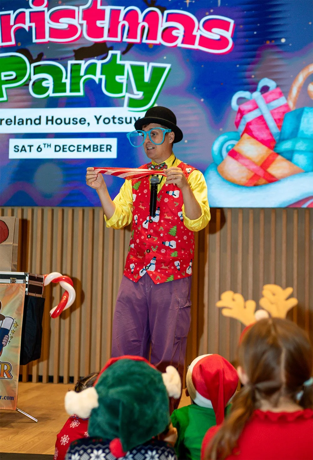 wearing a black hat, large blue glasses, a yellow shirt, and a red vest with holiday-themed patterns, standing on stage and holding a red and white striped ribbon in front of a large Christmas party advertisement. children in holiday costumes are sitting in front of the stage.