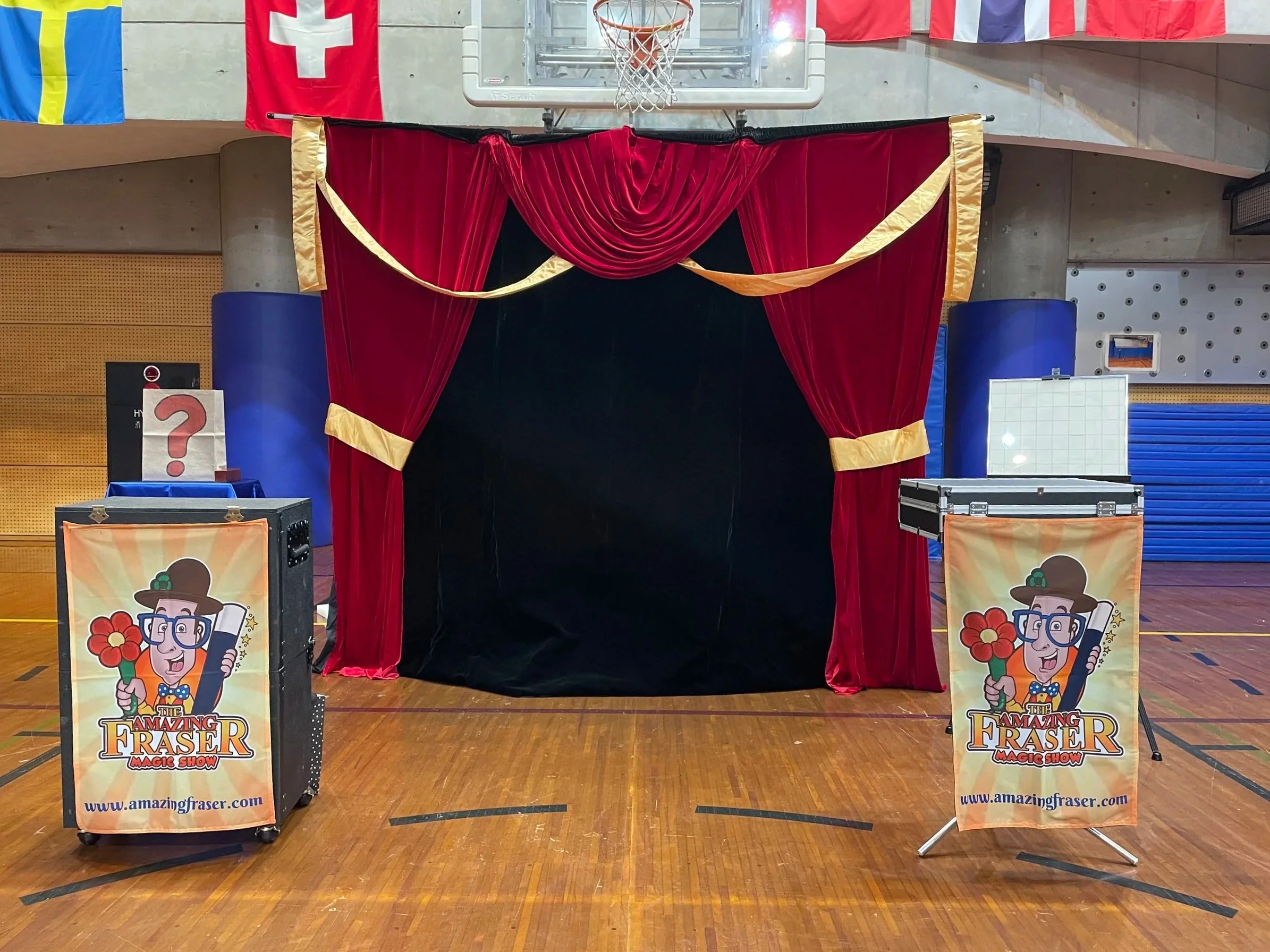 Empty stage with red velvet curtains and double yellow trim, set up for a magic show. A basketball hoop and flags are visible in the background. Two signs promoting the 'Amazing Fraser' magic show are positioned in front of the stage.