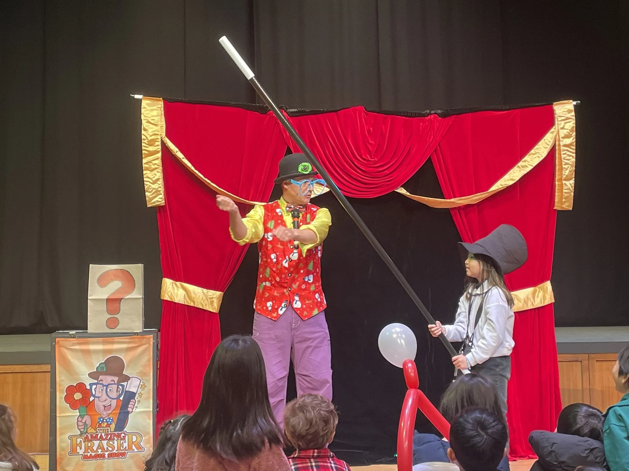 A performer dressed in colorful, whimsical clothing with glasses, a hat, and a headlamp, is on stage entertaining children. Behind him is a red curtain with gold trim, and a young girl wearing a hat and gray jacket is standing nearby. Audience member