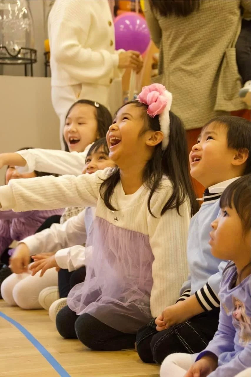 Children sitting on the floor, happily laughing and reaching out, likely at a children's event or party.