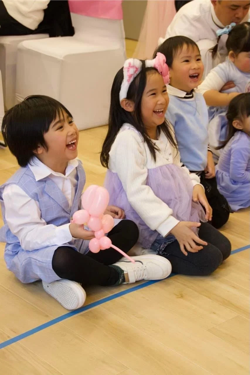 Group of children sitting on a wooden floor, laughing and smiling, with a girl holding a pink balloon animal, in a festive indoor setting.