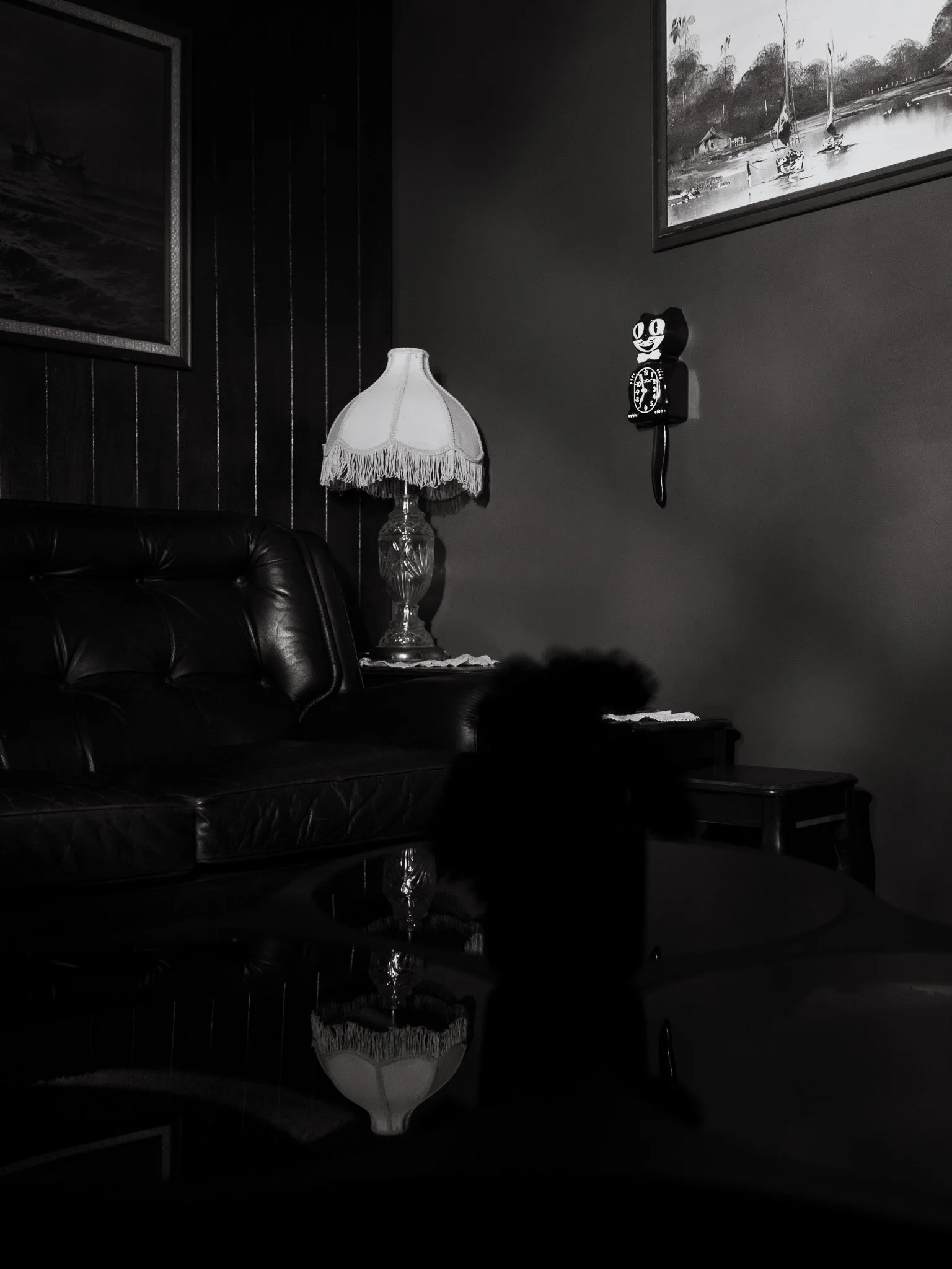 A black and white room with framed paintings on the walls, a vintage lamp on a table, a leather sofa, a small side table, and a decorative clock on the wall.
