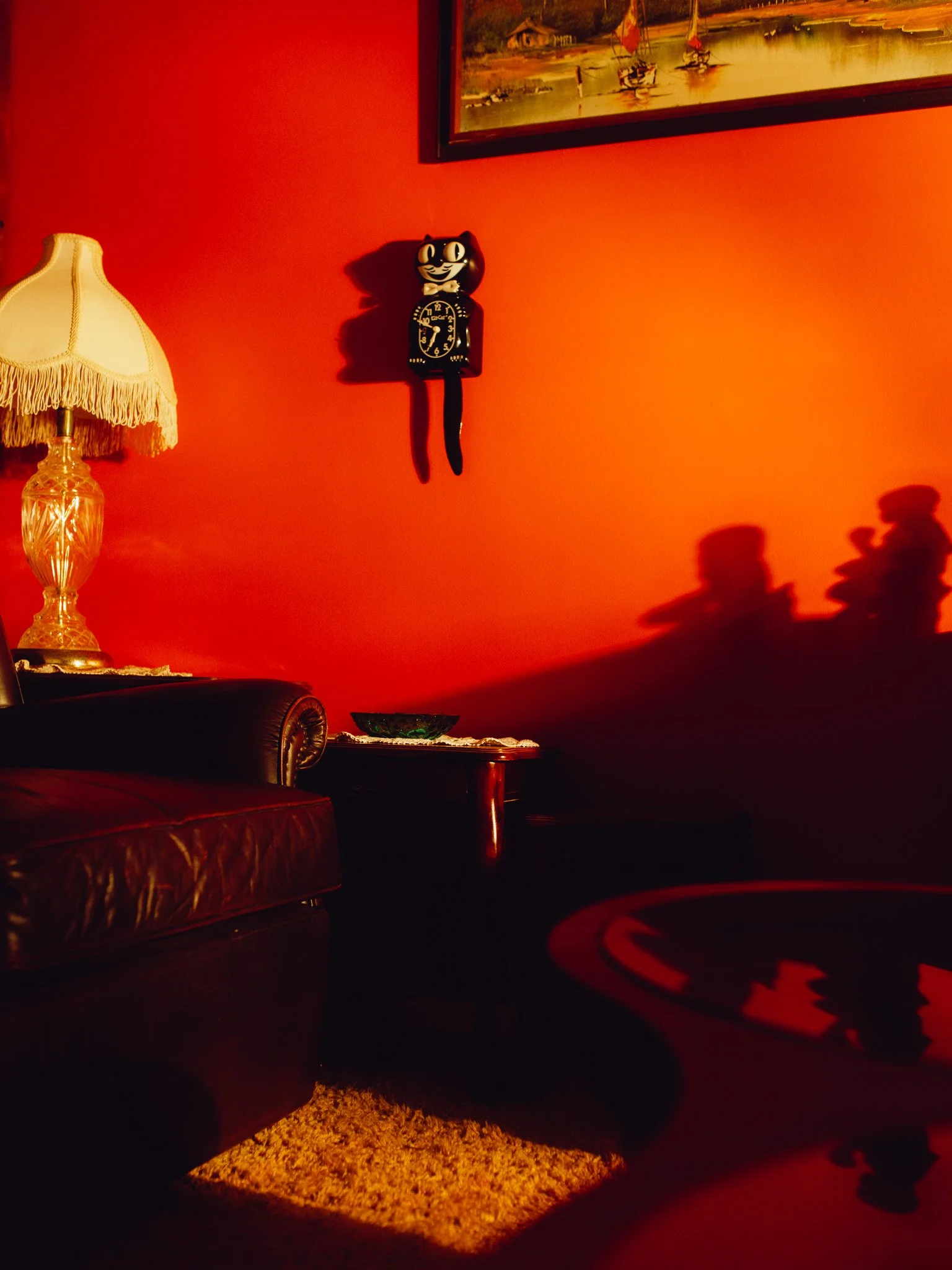 A dimly lit room with a red wall, featuring a vintage table lamp with a fringed lampshade, a small side table with a decorative bowl, a wall clock shaped like a black cat with its tail hanging down, a painting depicting boats on water, and the shadows of a teddy bear and a small figure cast on the wall.
