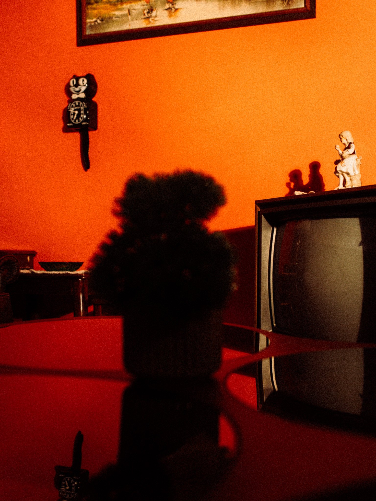 A silhouette of a person in front of a red wall with various decorations. On the wall, there is a black clock with a cat figure on top and a framed picture above it. To the right, there is a white figurine of a girl sitting and holding an object, casting shadows on the wall. There is an old television set with a curved screen on the right side of the image.