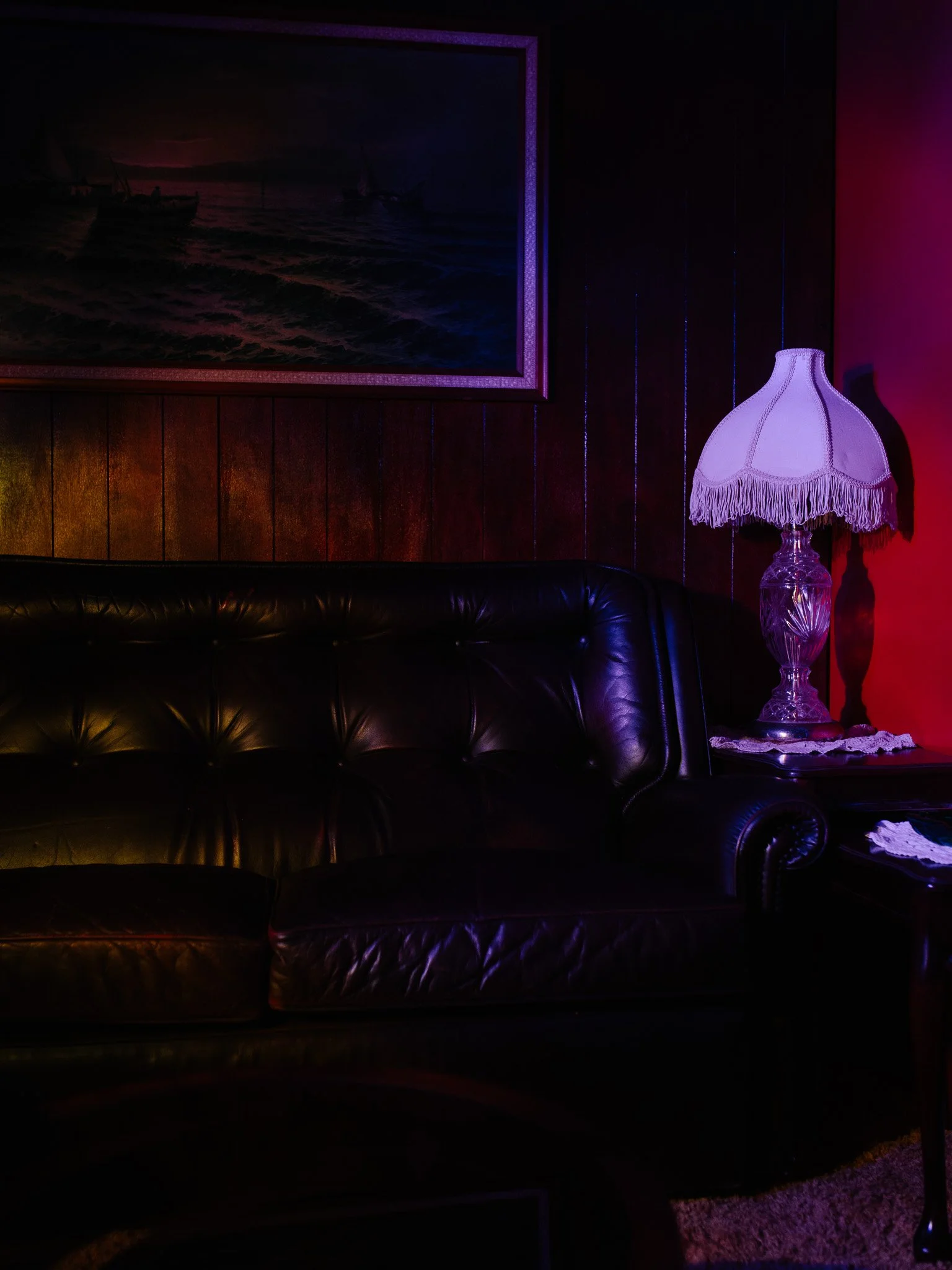 A dark living room scene with a black leather couch, a vintage glass table lamp with a fringed lampshade, a wood-paneled wall, a framed nautical painting, and some colored lighting reflections.