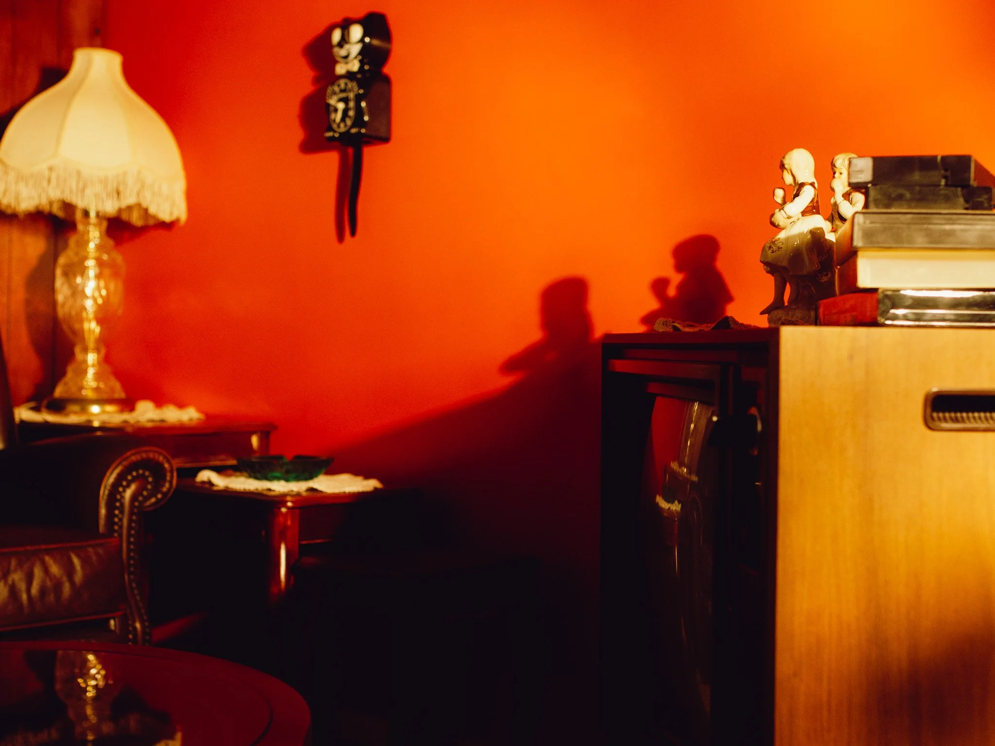 A corner of a room with a red wall, featuring a vintage table lamp with a fringed lampshade on a side table, and decorative figurines of two women sitting on a bookend on a wooden cabinet, with a stack of books nearby.