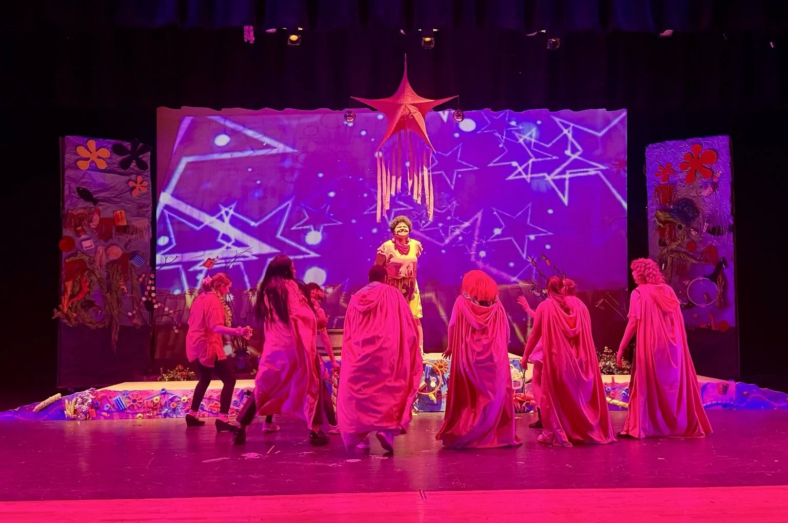 Student ensemble performing onstage beneath colorful theatrical lighting and projections.