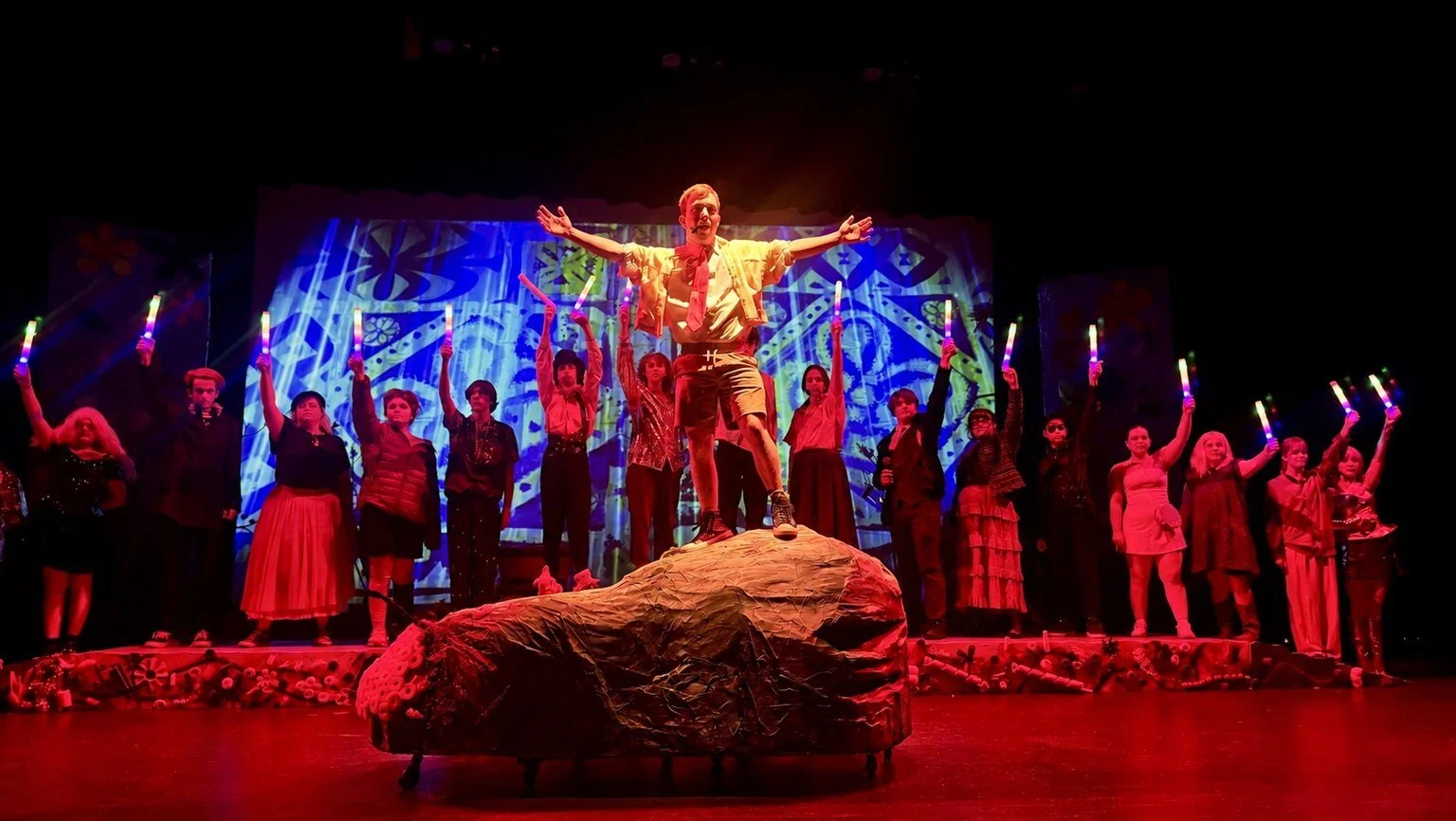 Student theatre ensemble performing onstage under dramatic red lighting, with a performer standing on a large central scenic rock.