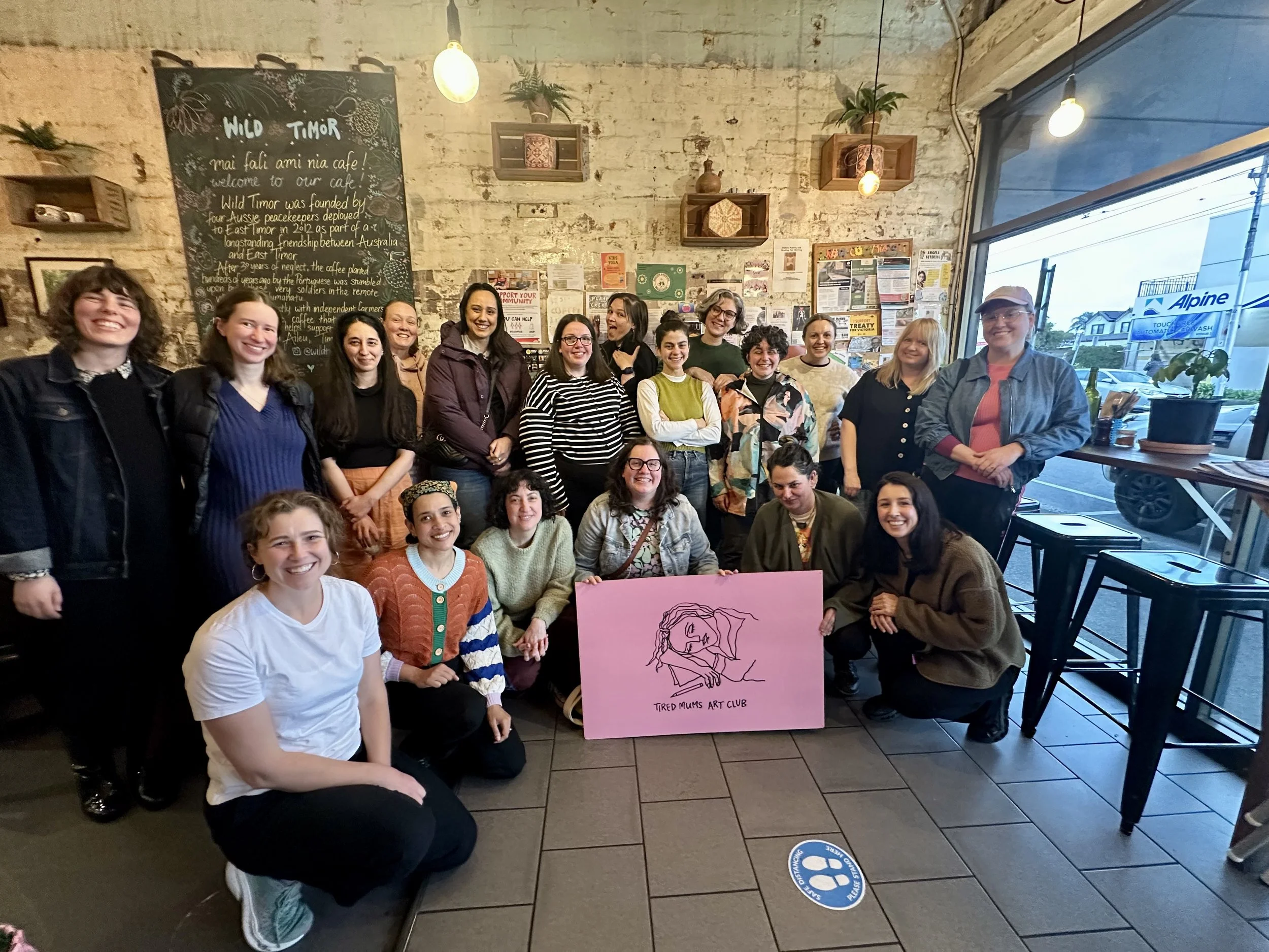 A group of 14 women and girls gathered in a cafe, smiling for the camera. They are standing and sitting in front of a wall decorated with a chalkboard, shelves with plants and decorative items, and various posters. A pink sign with an illustration and the text 'TIRED MUMS ART CLUB' is held by one of the women in the front, and there is a large window with a street view on the right.