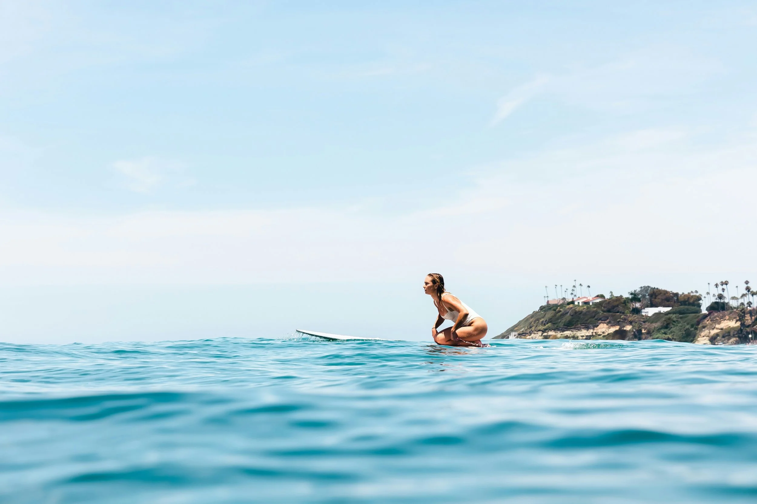 A woman kneeling on a surfboard in the ocean, with a coastal hillside and houses in the background, under a clear sky.