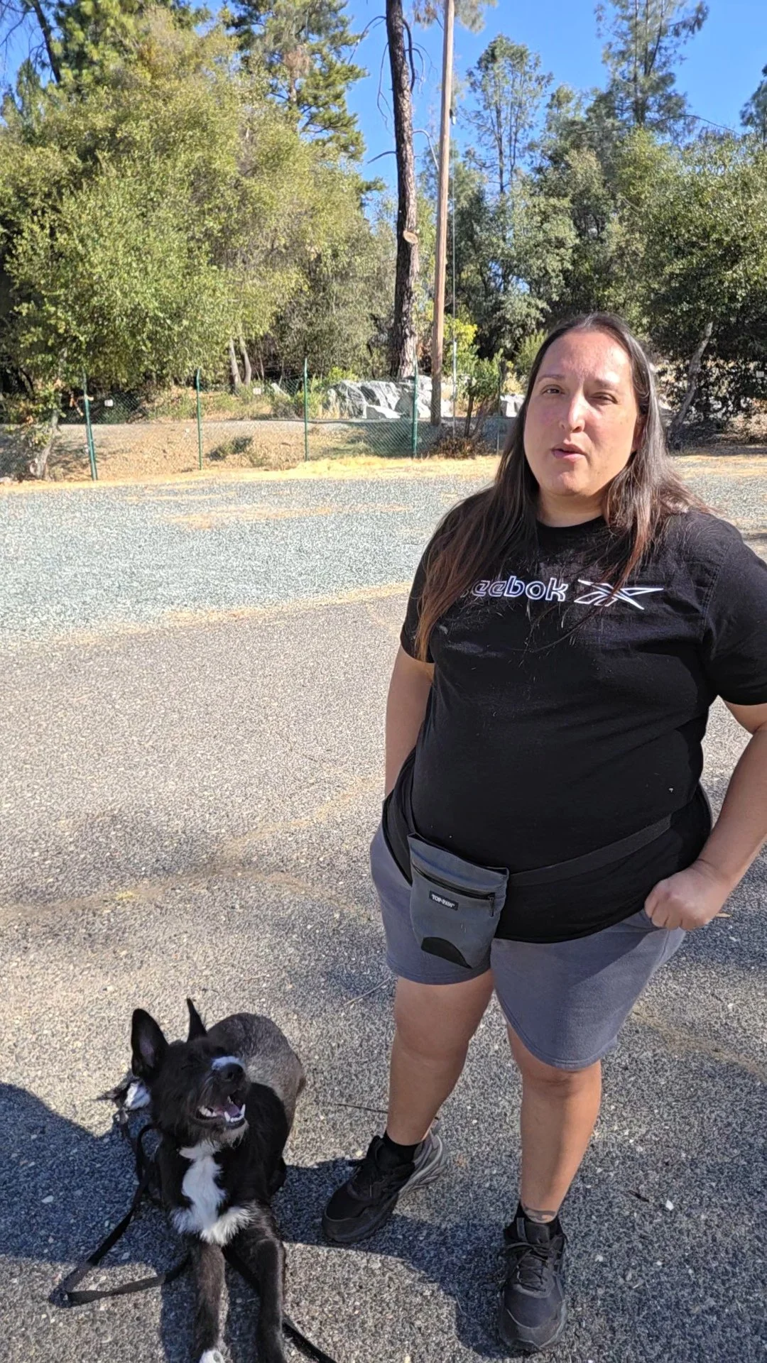 A woman standing on a paved outdoor area with a black and white dog, trees, and a fence in the background on a sunny day.