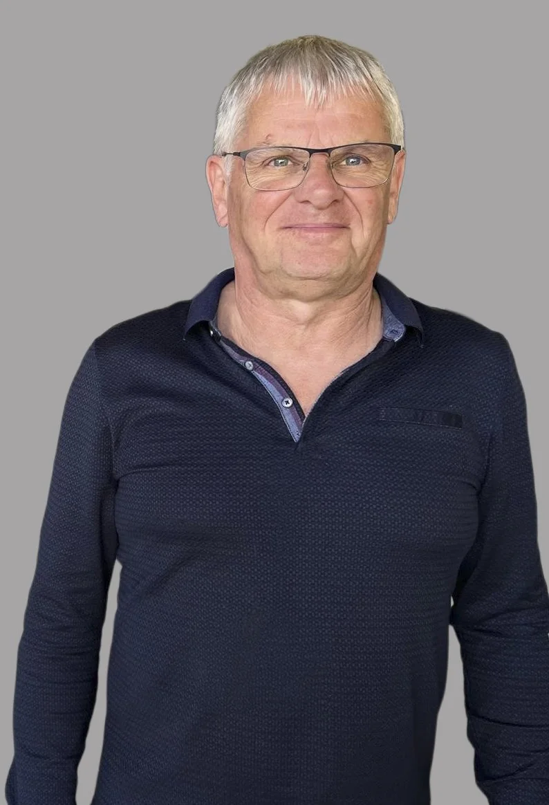 Homme âgé portant des lunettes, souriant, en chemise à col et pull sombre, fond gris.