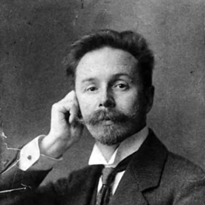 Black and white portrait of a man with a beard and mustache, dressed in late 19th or early 20th-century attire, resting his head on his hand.