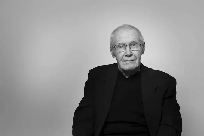 Black and white photo of an elderly man with glasses, wearing a dark blazer and shirt, sitting against a plain background.