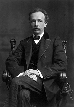 Black and white portrait of a man in formal attire seated in a wooden chair, wearing a suit, bow tie, and vest with a serious expression.
