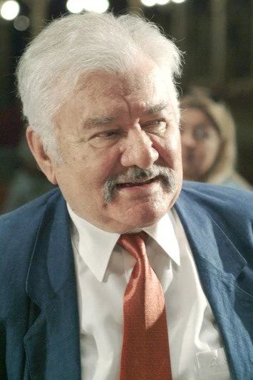 Close-up of an older man with gray hair and a mustache, wearing a white shirt, a red tie, and a blue blazer, in an indoor setting.