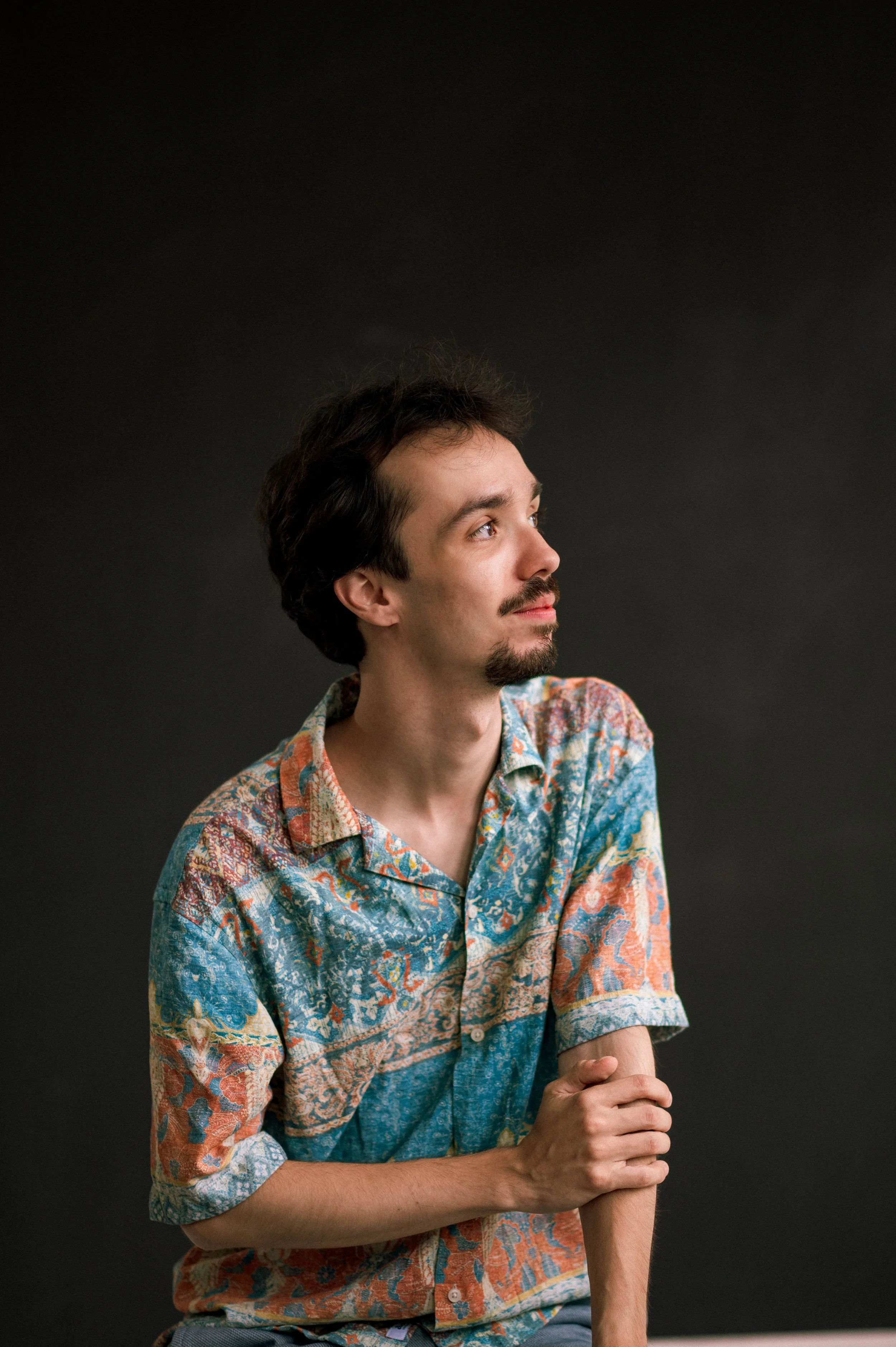A young man with dark hair and a goatee, dressed in a colorful, patterned short-sleeve shirt, sits against a black background. He is looking to his right with a contemplative expression and his hands clasped together on his lap.