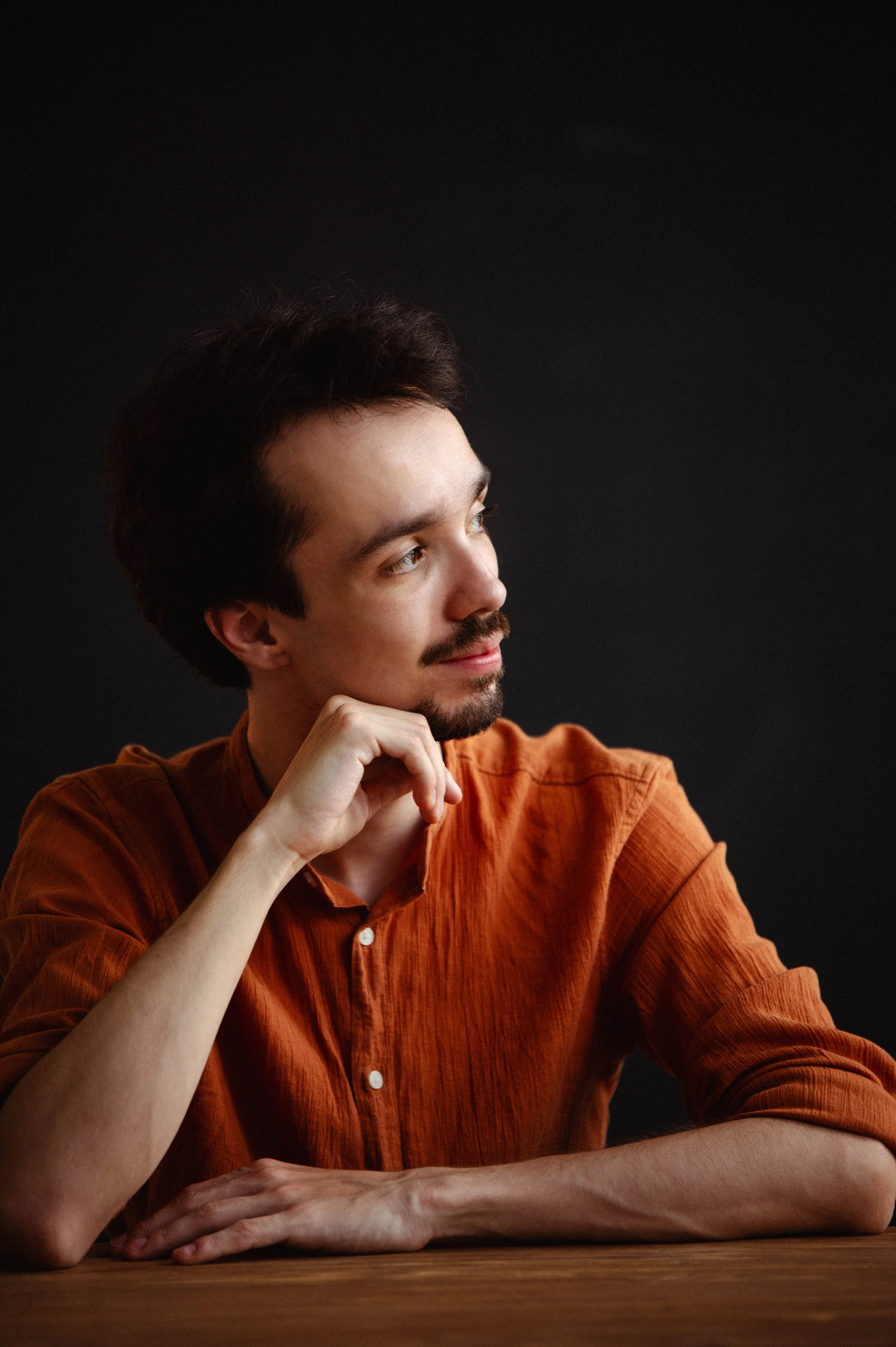 A young man with dark hair and a beard, wearing an orange shirt, sitting at a wooden table, resting his chin on his hand, and looking thoughtfully to his left against a black background.