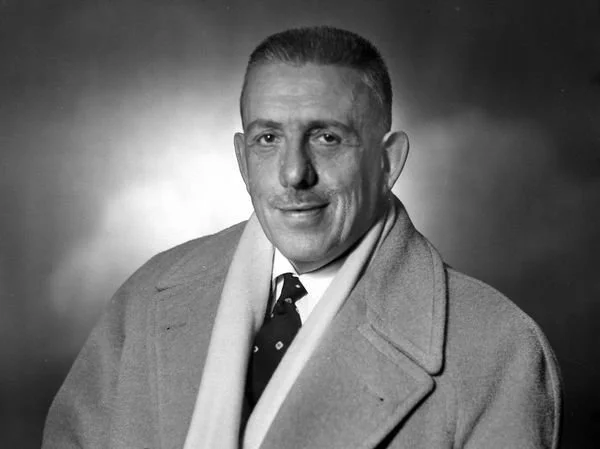 Black and white portrait of a man wearing a coat and tie, smiling slightly with short, neatly combed hair.
