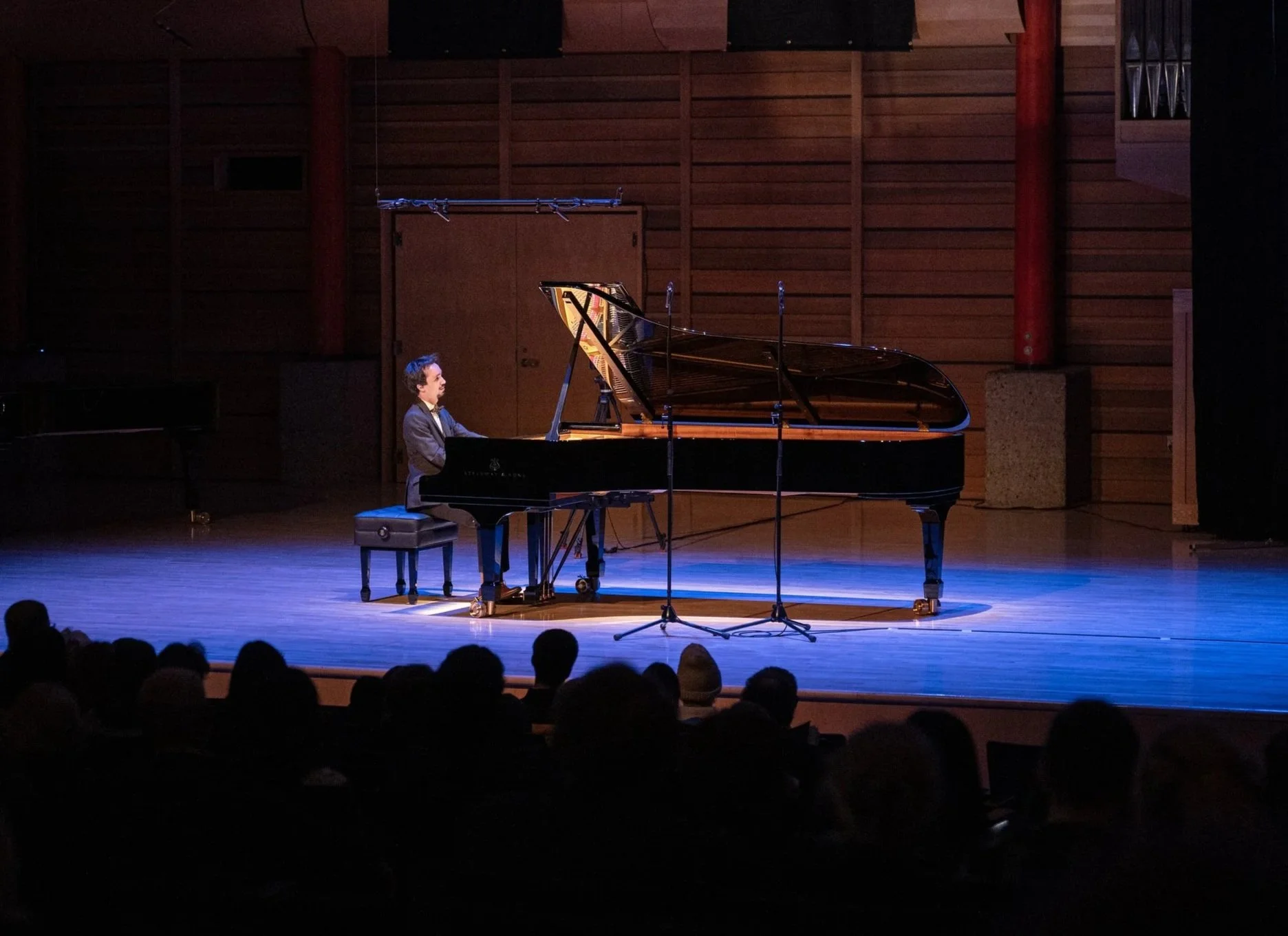 A pianist performing on stage in a concert hall, with a grand piano and microphones, and an audience watching.