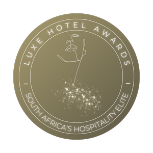 Gold circular badge with the text 'LUXE HOTEL AWARDS' and 'SOUTH AFRICA'S HOSPITALITY ELITE' around the border. In the center, a stylized outline of a trophy or cup with a flag on top, and a sparkling trail beneath it.
