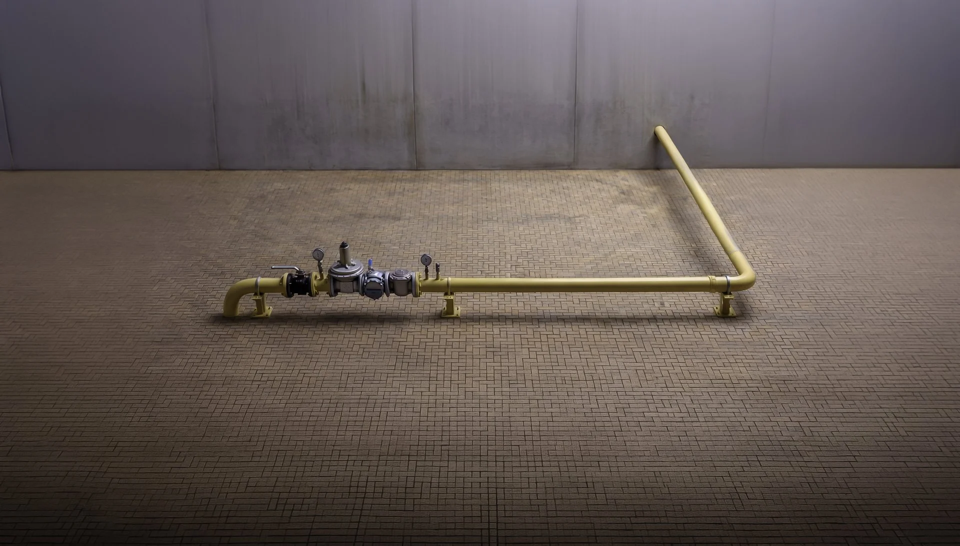 A yellow pipeline with gauges and valves on a brick floor, against a metal wall background.