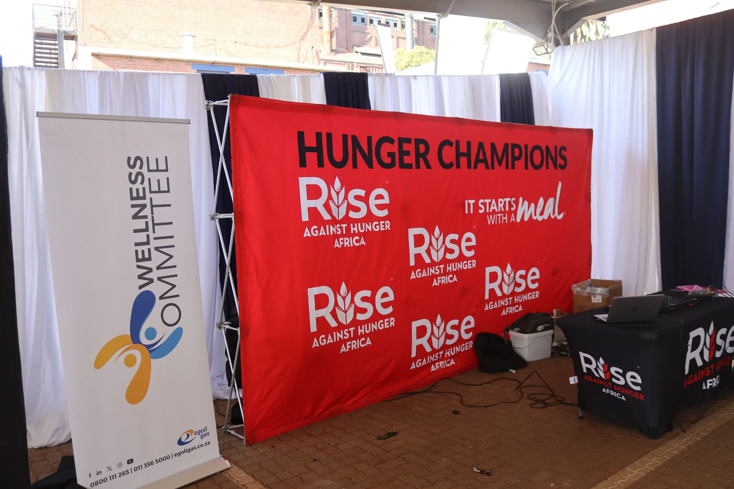 An outdoor display booth with banners and tables promoting Hunger Champions Rise Against Hunger Africa. The booth has a red banner with the organization's name and tagline, set against a backdrop of white, blue, and dark-colored curtains.