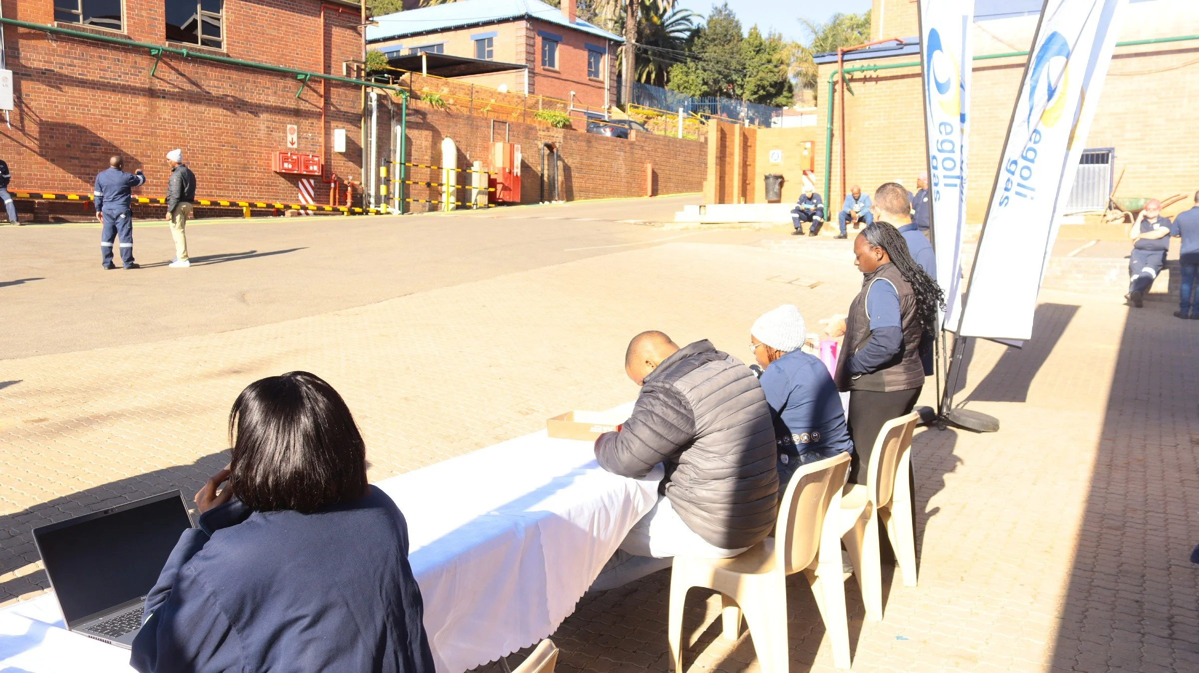 People seated at tables and standing outside, with a few cars and a brick building in the background. It appears to be an outdoor community or office event on a sunny day.