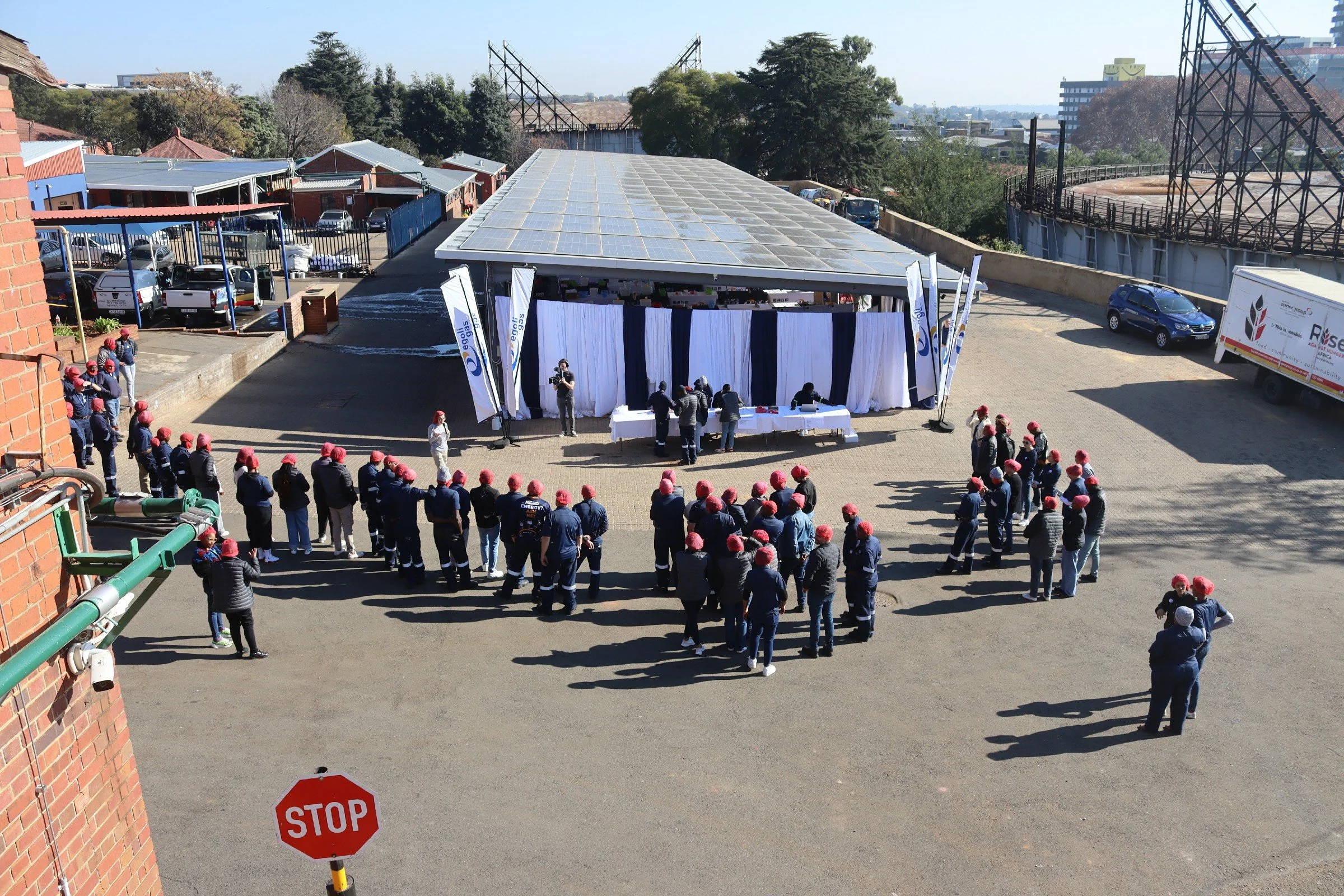 A gathering of people wearing red hats and blue uniforms standing outside a building with a large covered area with solar panels on the roof. There are banners and a table set up in front of the covered area, and several people are talking near the t