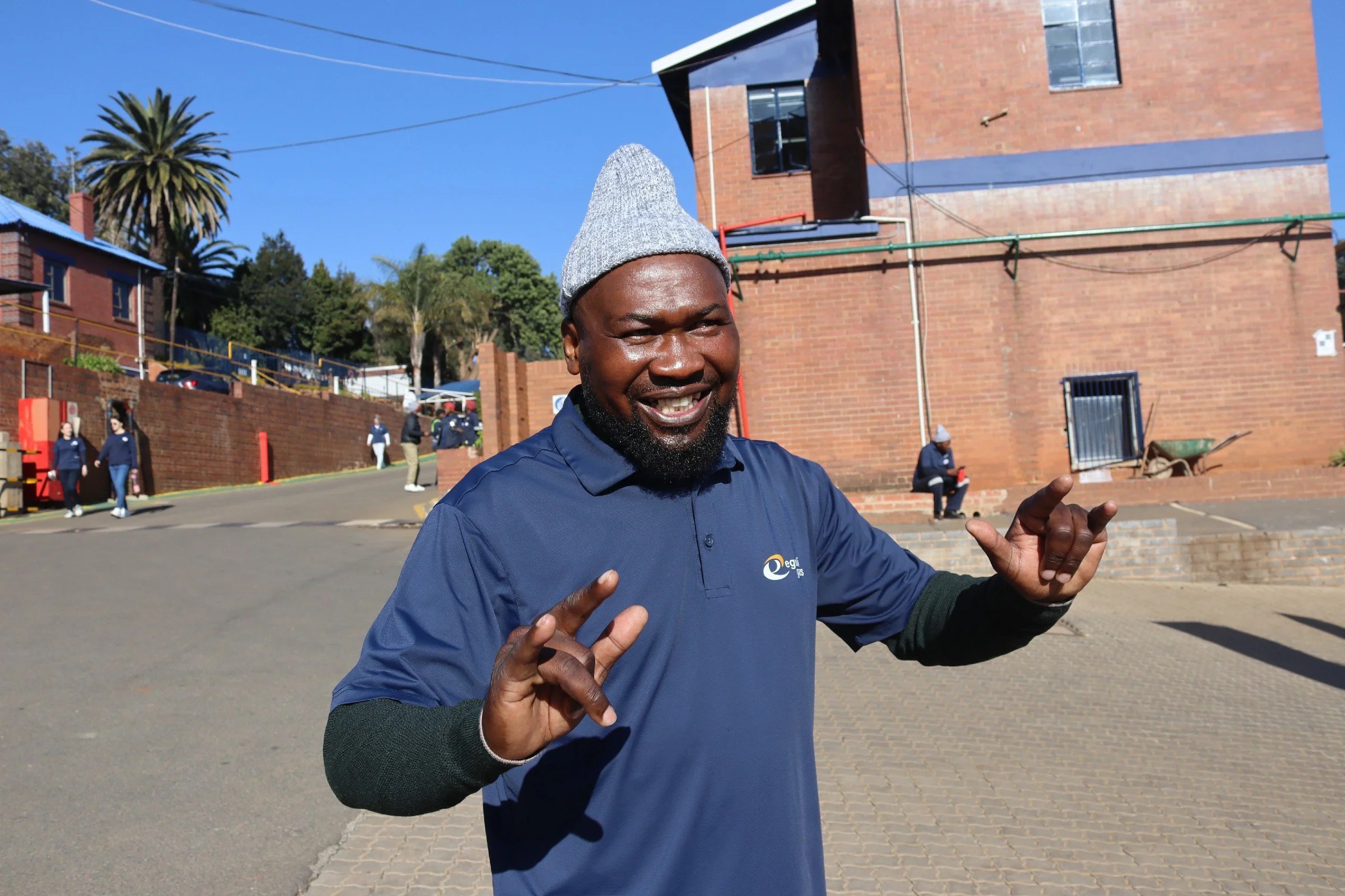 A man with a beard wearing a gray knit beanie and a blue polo shirt, making hand gestures while smiling outdoors on a sunny day, with a brick building and other people in the background.