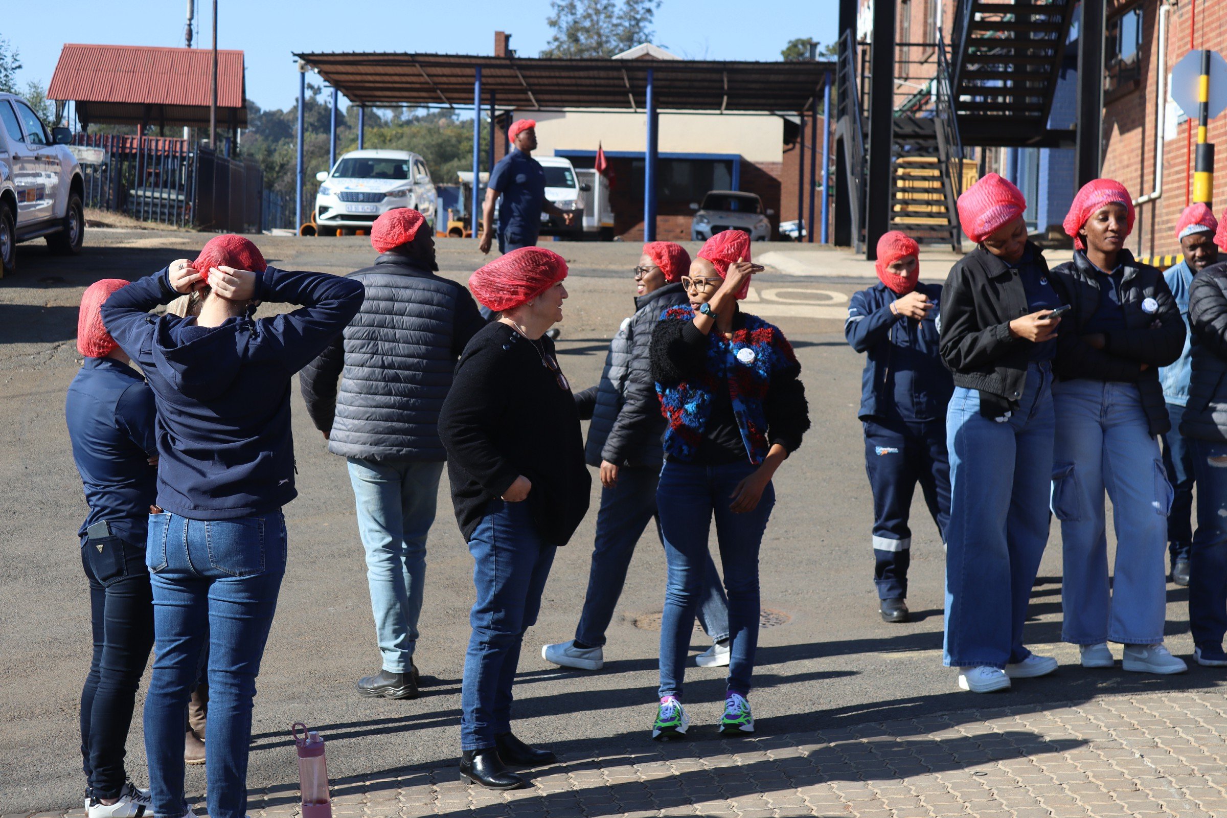 A group of people, mostly women, wearing red hair nets, standing outdoors on a sunny day, some talking, others looking at their phones, with parked cars and buildings in the background.