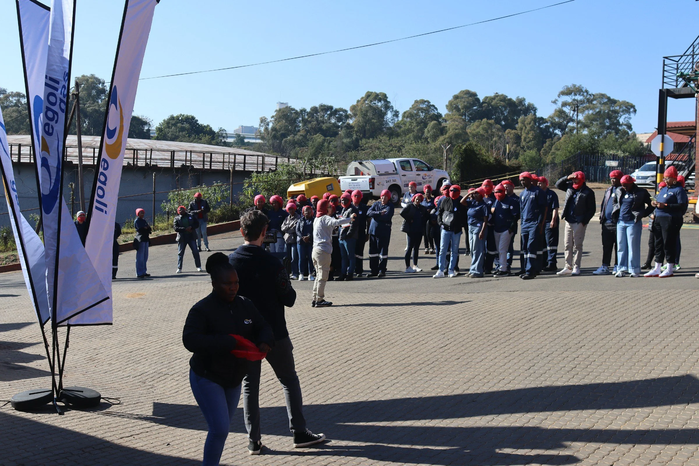 Group of people standing outdoors on a sunny day, some wearing red head coverings, with flags bearing a logo, and a person gesturing while others watch. There are trees and vehicles in the background.