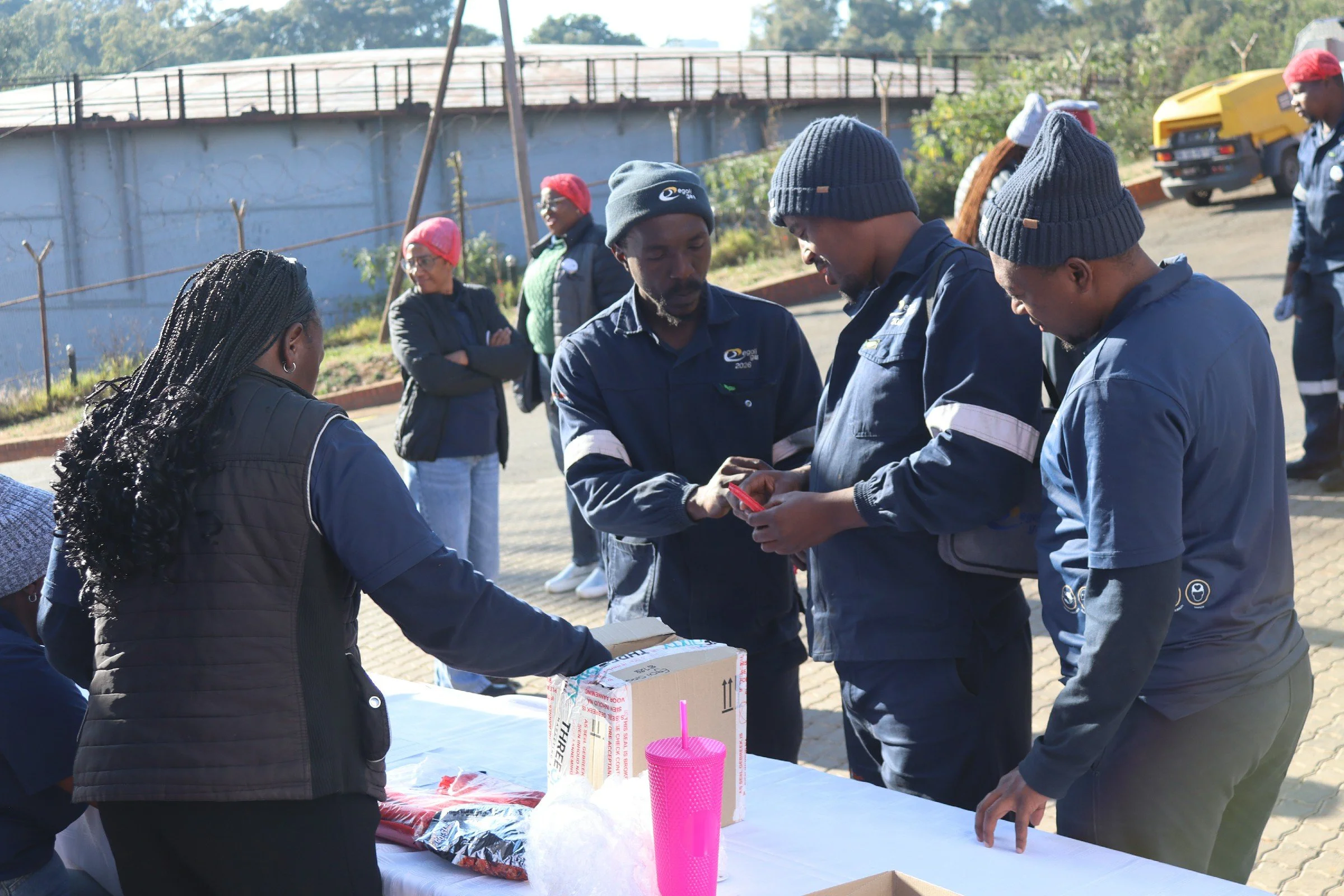 People wearing uniforms gathered around a table outdoors, some looking at items and others engaged in conversation, with a fence and trees in the background.