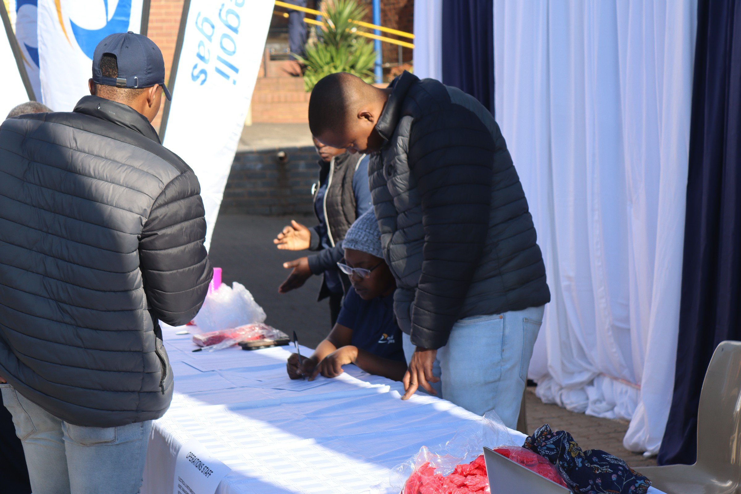 Group of people at an outdoor table, some signing papers, at a community event. There are banners and a white curtain in the background.