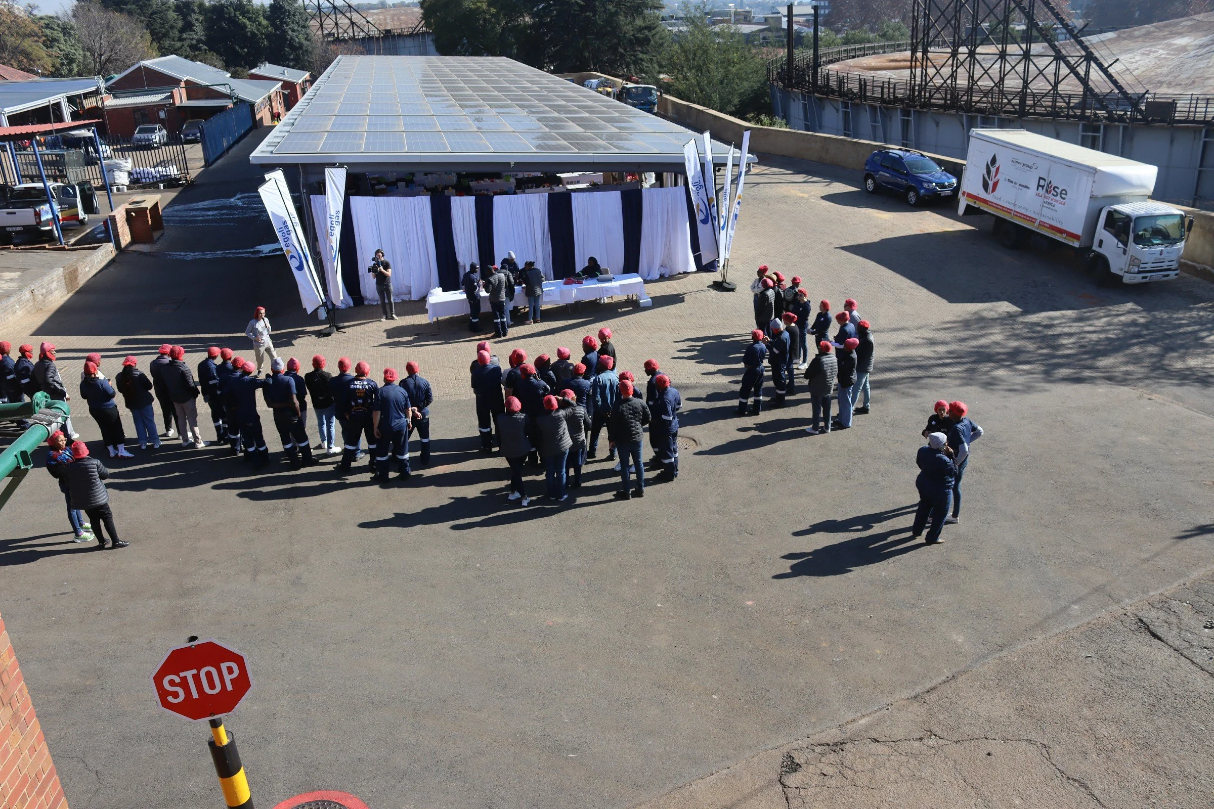People gathered outdoors for an event, with a stage decorated with flags and white curtains. All attendees are wearing red hats and are standing in front of the stage. There is a white truck and a car parked nearby, and a stop sign is visible in the 