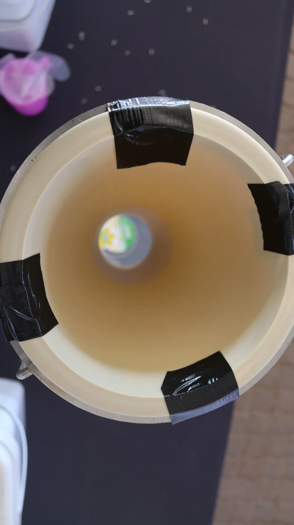 A top view of a hollow, beige-colored object with black tape on the rim, placed on a dark surface. In the background, there is a blurred purple and white object and some scattered small dots.