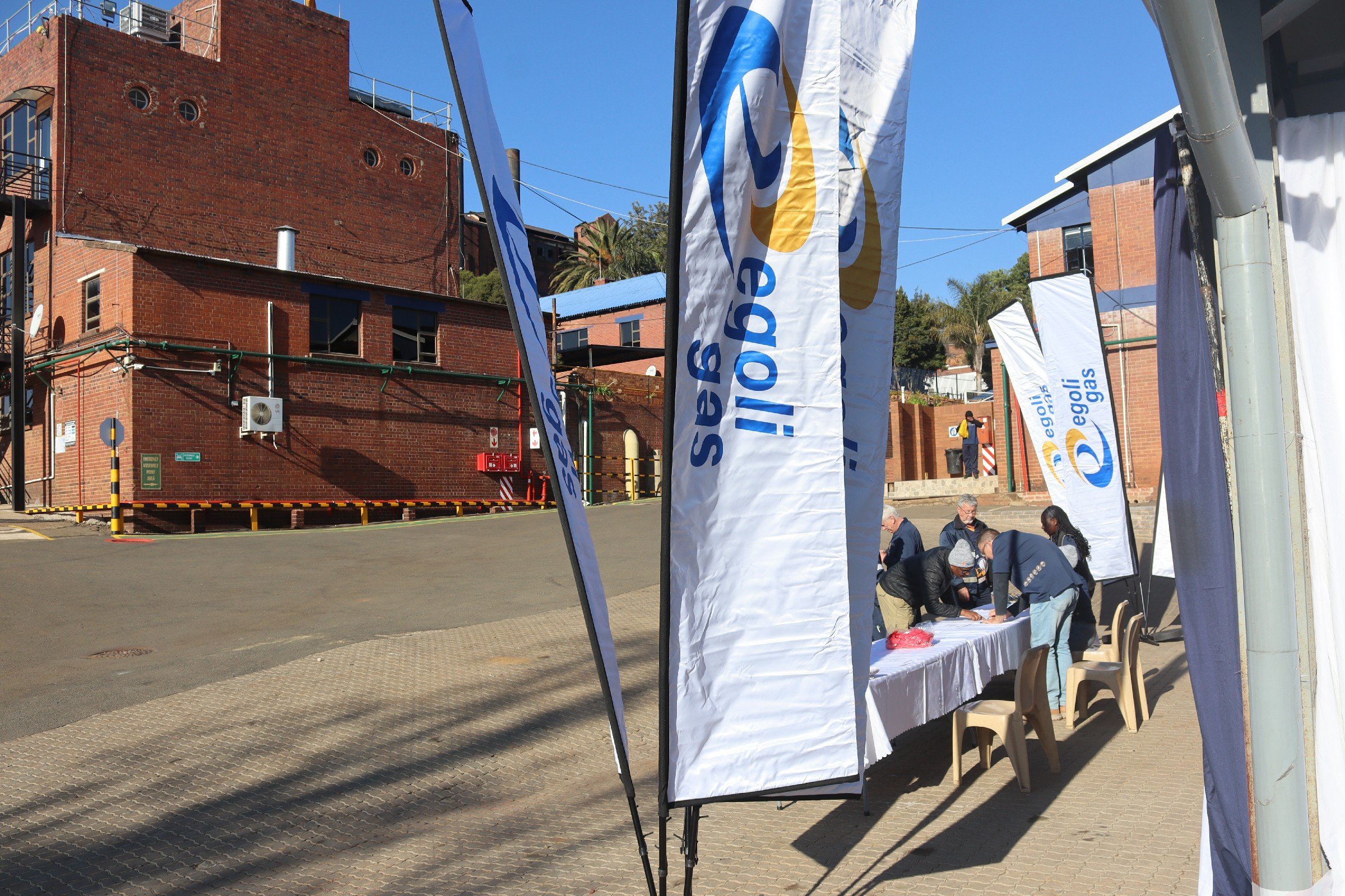 Outdoor scene with American Flags and banners that read 'Egas' and 'Eggs', with a table where people are gathered, and brick buildings in the background under a clear blue sky.